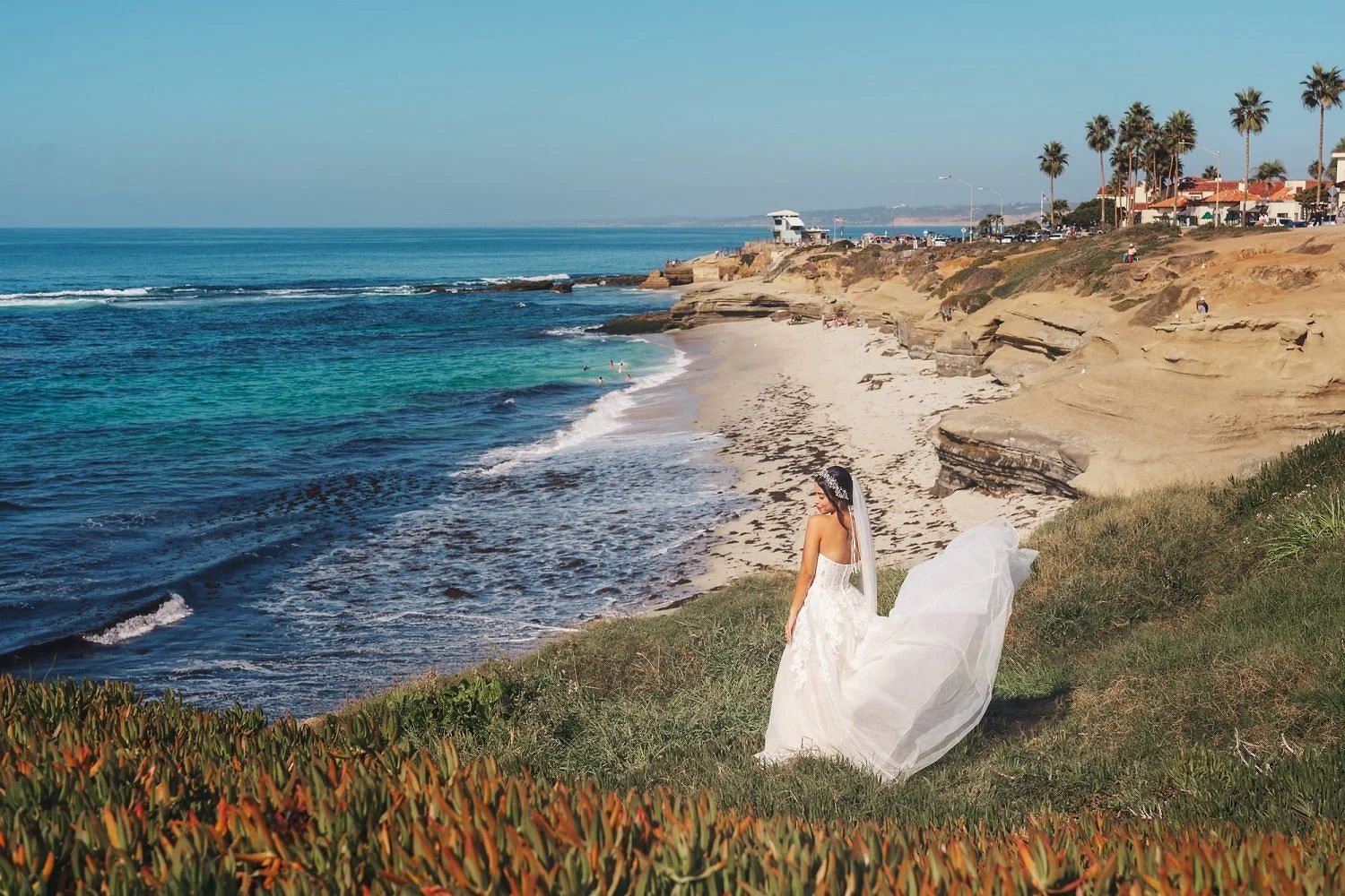 A woman in a wedding dress standing on grassy area overlooking the ocean beach with rocky cliffs, palm trees, and buildings in the background.