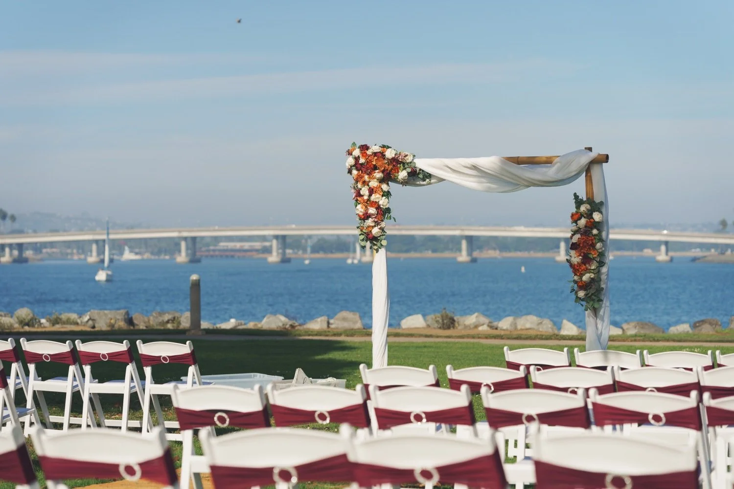 Outdoor wedding setup near water with rows of white chairs with red sashes and a decorated arch with flowers and white fabric, overlooking a bridge and sailboats.