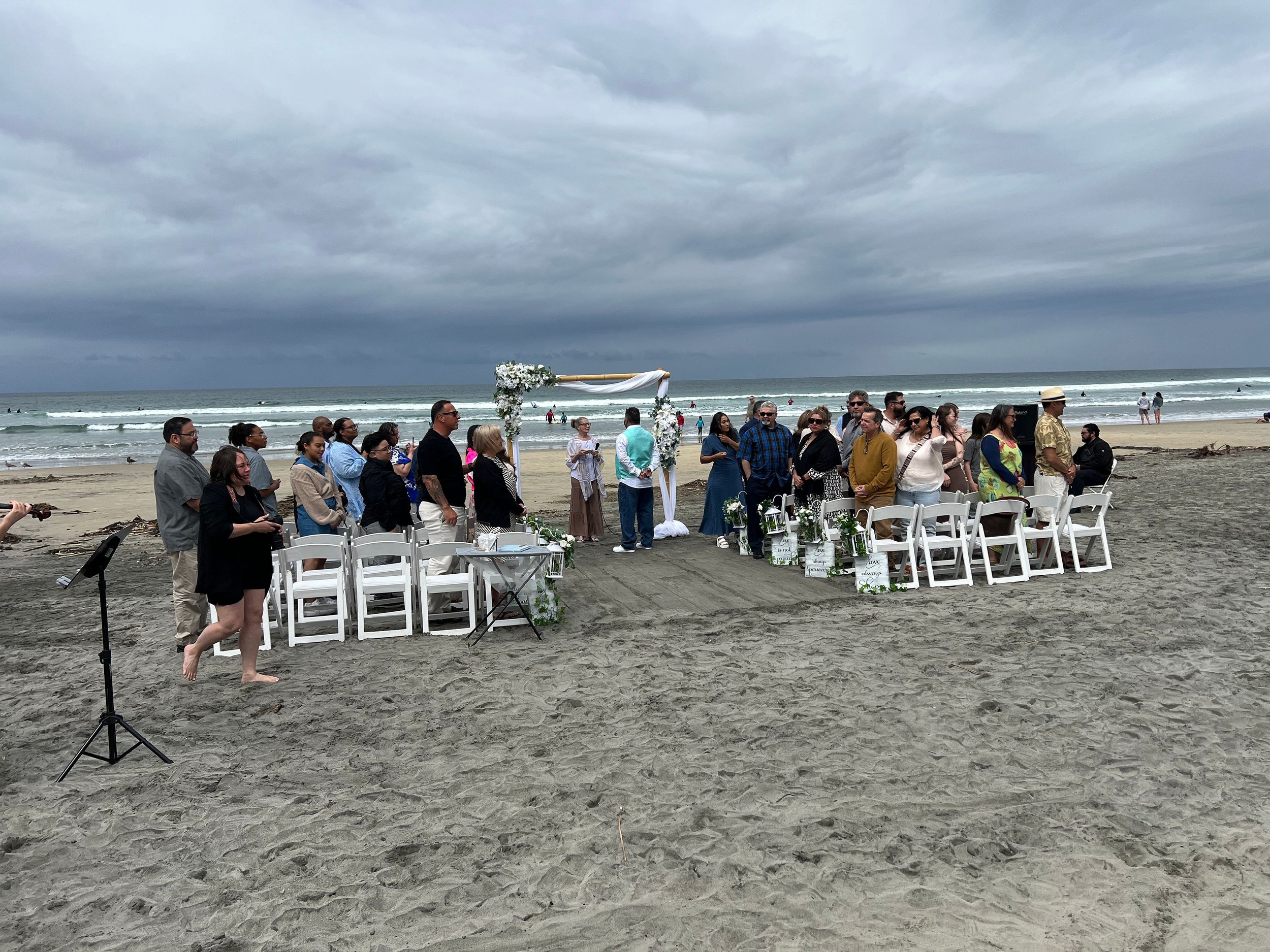 A group of people gathered on a beach for a wedding ceremony under dark cloudy skies, with chairs, floral decorations, and an archway.
