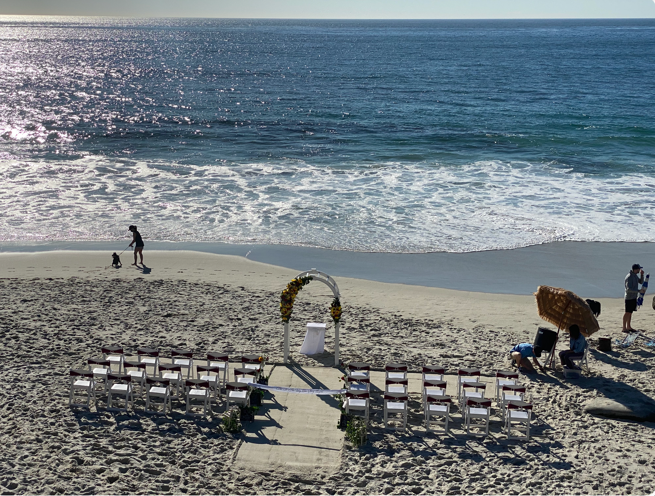 A beach scene with rows of white chairs set up for a wedding, facing the ocean. People are preparing, with some arranging decorations and others holding umbrellas. The ocean is sparkling in the sunlight, with waves gently rolling onto the shore.