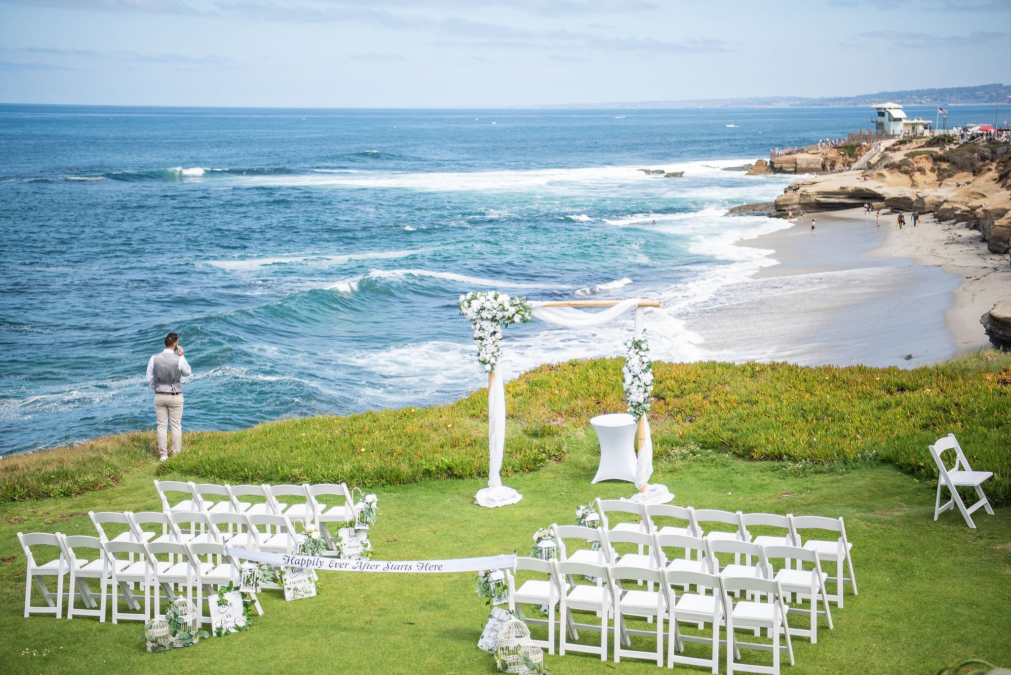 A wedding ceremony setup on a grassy hill overlooking the ocean, with white chairs arranged in rows, a floral arch, and a man in a vest and pants standing on the grass near the water.