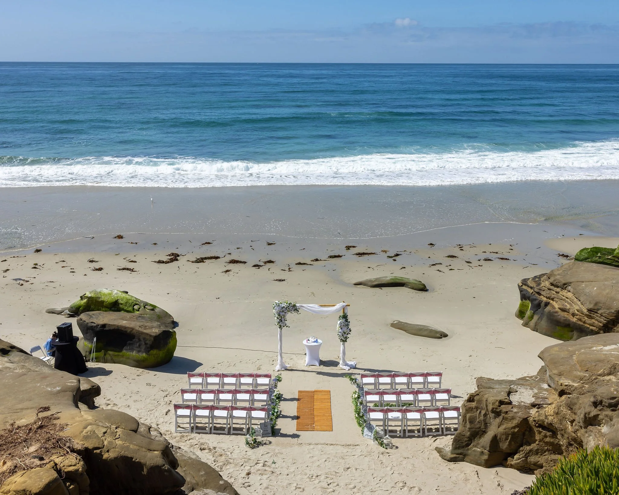 A wedding setup on the beach with rows of white chairs facing a small altar decorated with flowers, situated on the sand between large rocks, with the ocean and blue sky in the background.