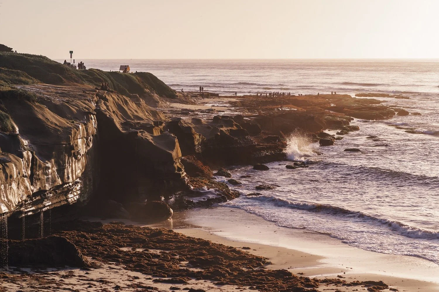 Coastal scene with rocky cliffs, sandy beach, and ocean waves at sunset, with people walking on the shore and atop the cliffs.