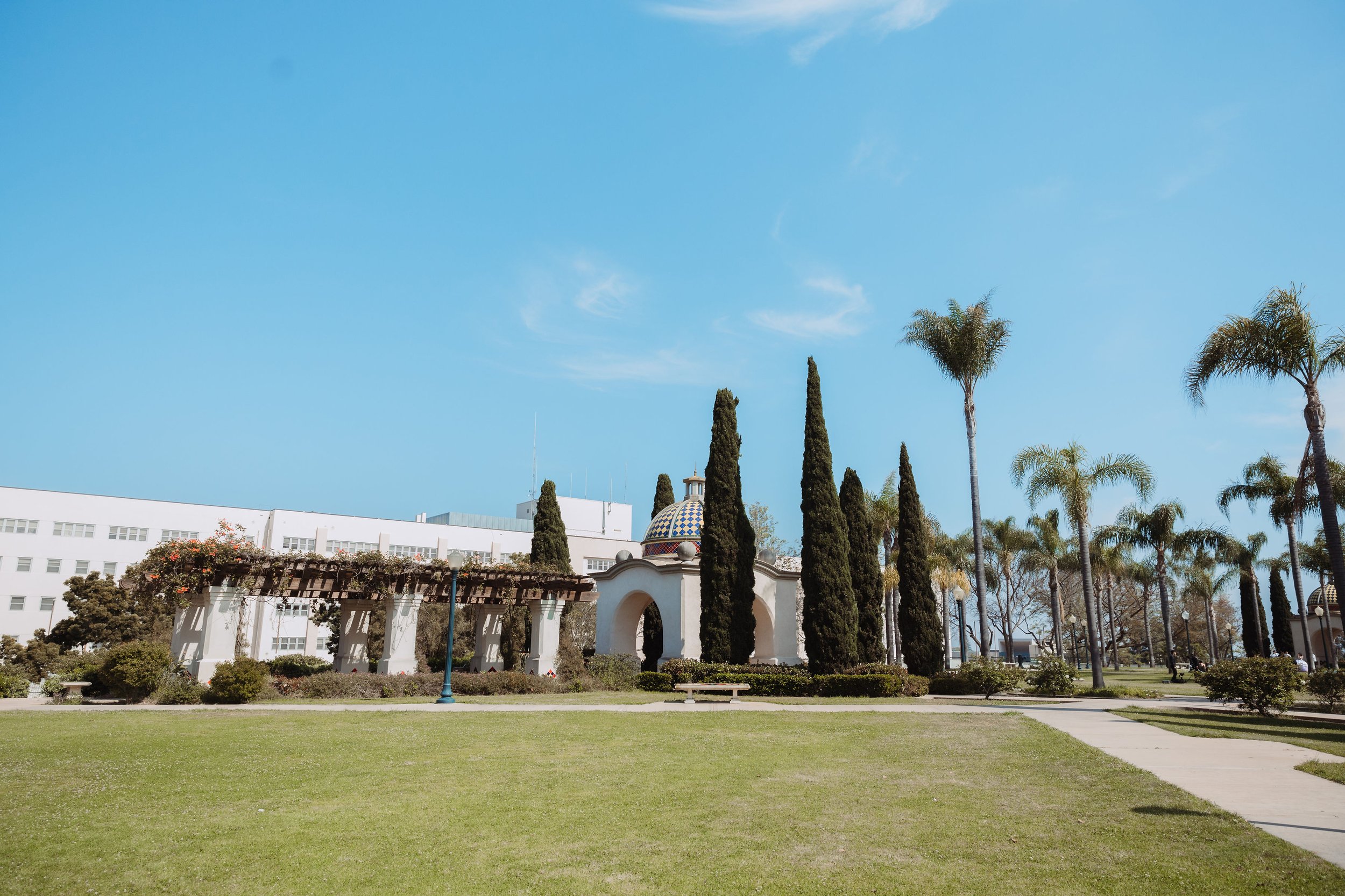 A park with green grass, a walkway, palm trees, cypress trees, benches, street lamps, and a building with white walls and a dome-shaped roof in the background.
