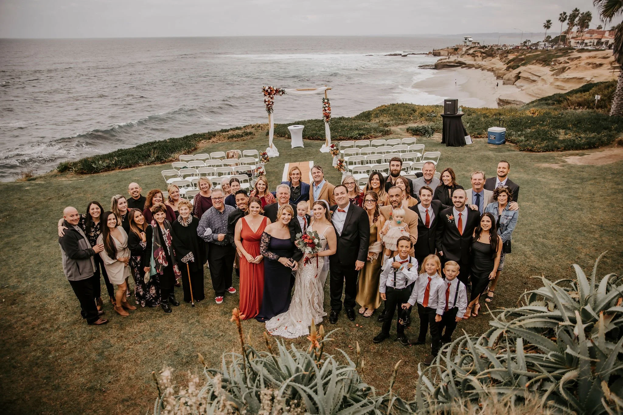 A group of people dressed for a wedding posing together on a grassy area near the beach with ocean waves, rocky cliffs, and buildings in the background.