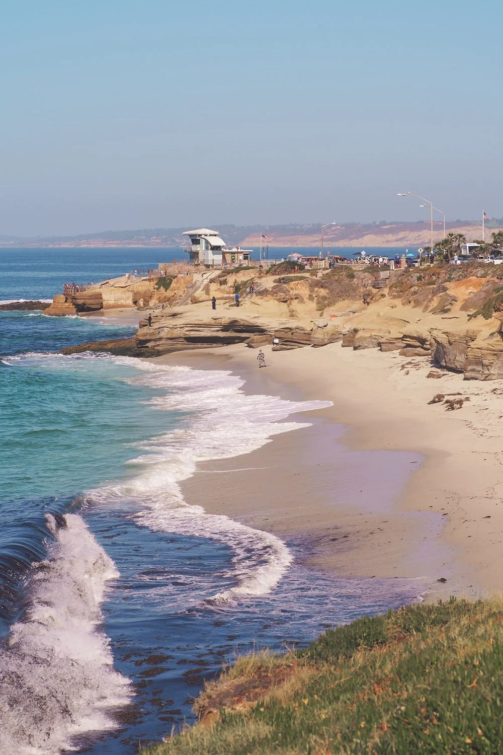 A sandy beach with waves crashing onto the shore, rocky cliffs in the background, and a lifeguard tower on the cliff overlooking the beach. There are people walking and relaxing near the water, with a clear blue sky above.