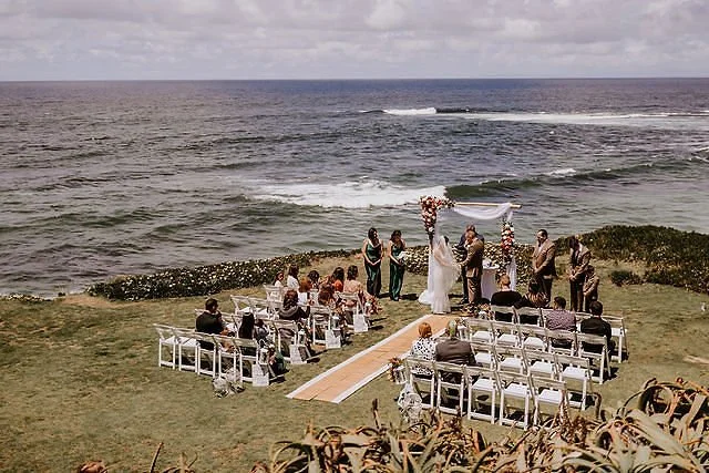 A wedding ceremony taking place on a grassy area by the ocean, with a couple exchanging vows under a decorated arch, surrounded by guests seated on white chairs.