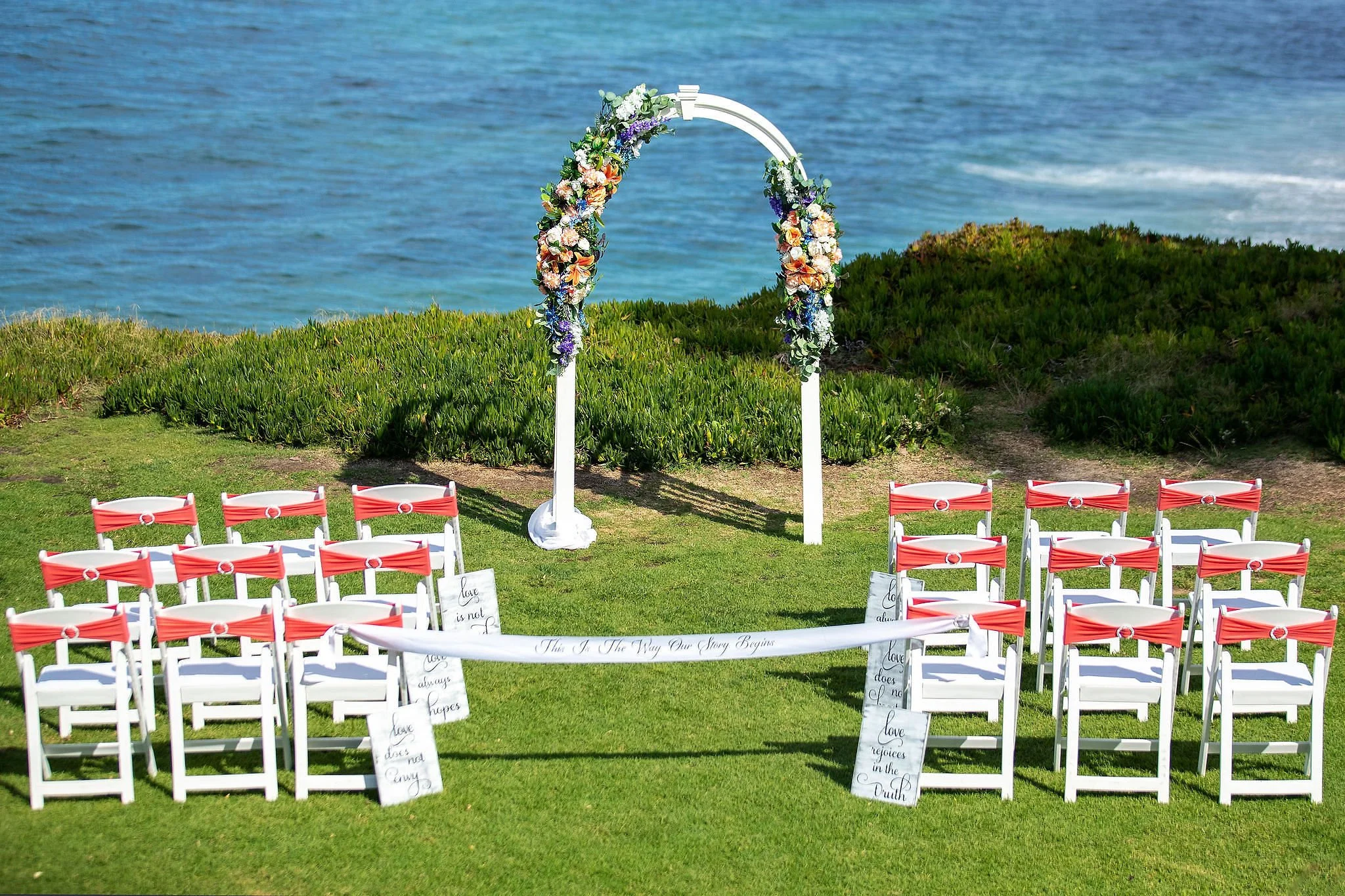 Outdoor wedding setup featuring rows of white chairs with red sashes, a floral wedding arch by the water, and decorative signs on the grass.