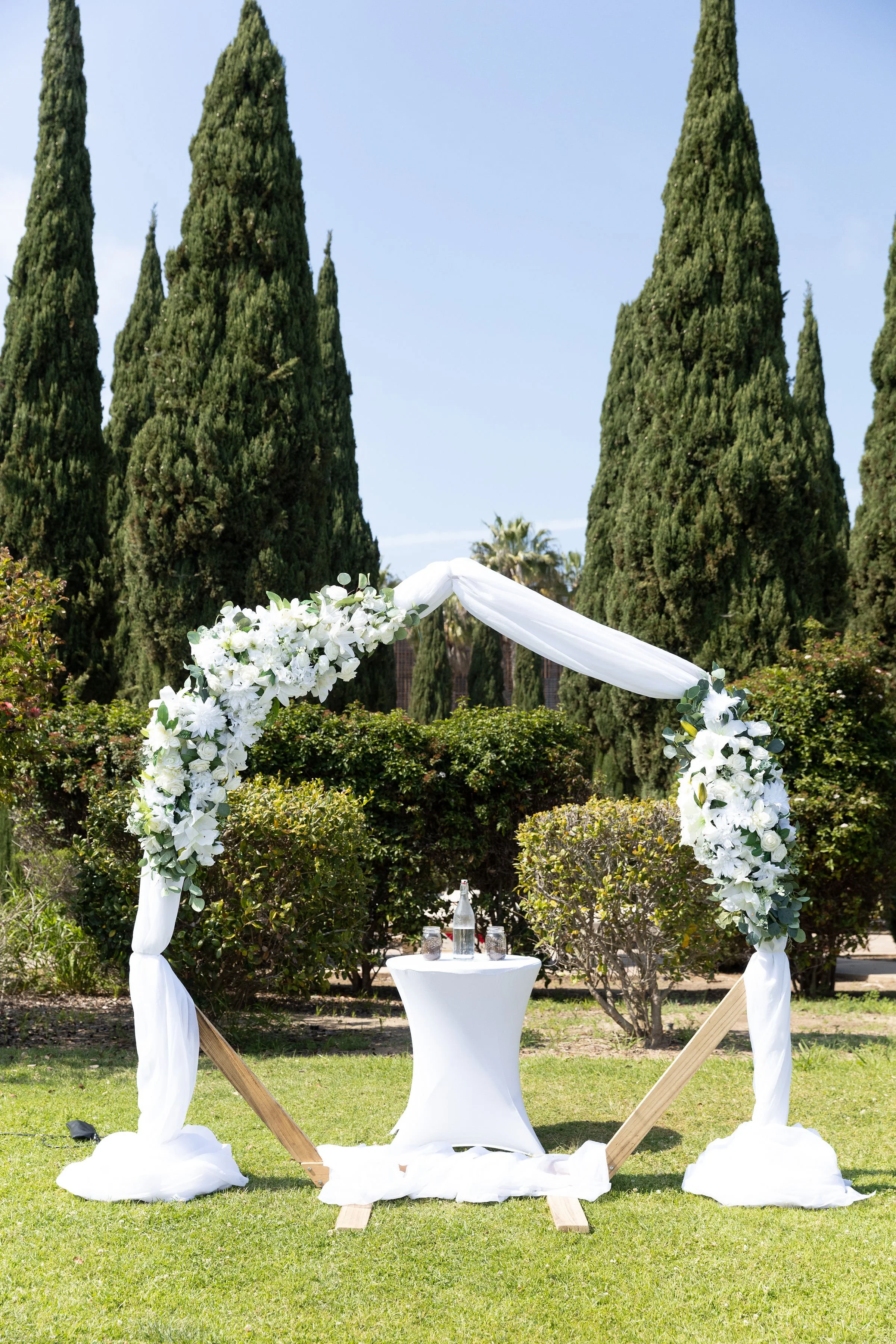 Wedding arch decorated with white flowers and white fabric, set up outdoors on green grass with trees in the background.