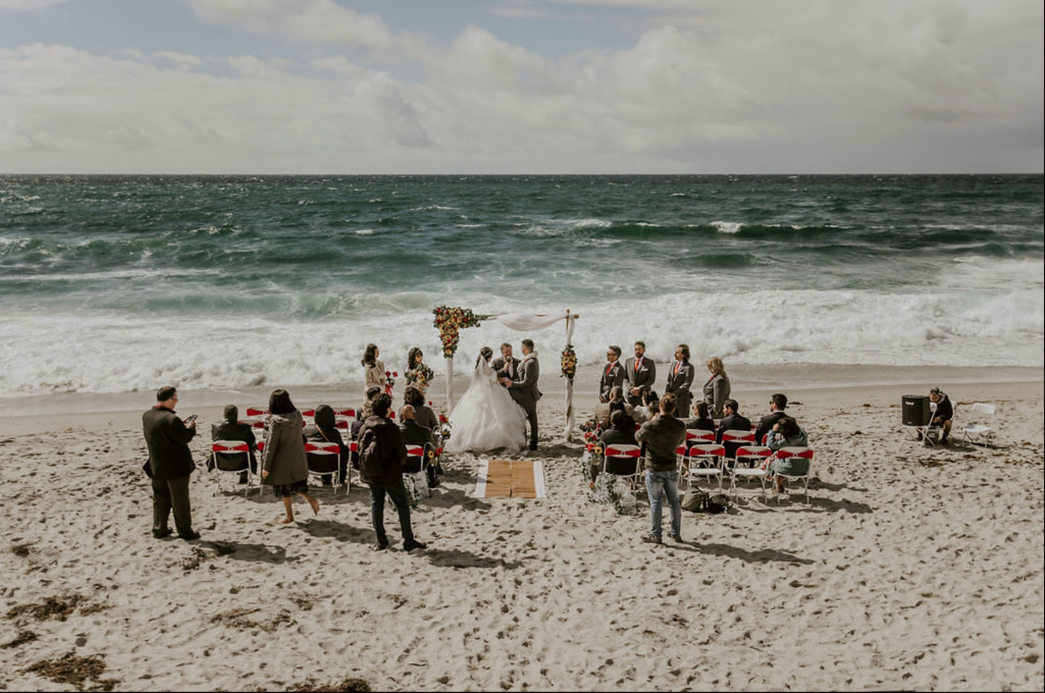 A wedding ceremony taking place on the beach with the ocean in the background. The bride and groom are standing under an arch decorated with flowers, surrounded by guests seated on chairs. The sky is cloudy, and people are dressed in formal attire.
