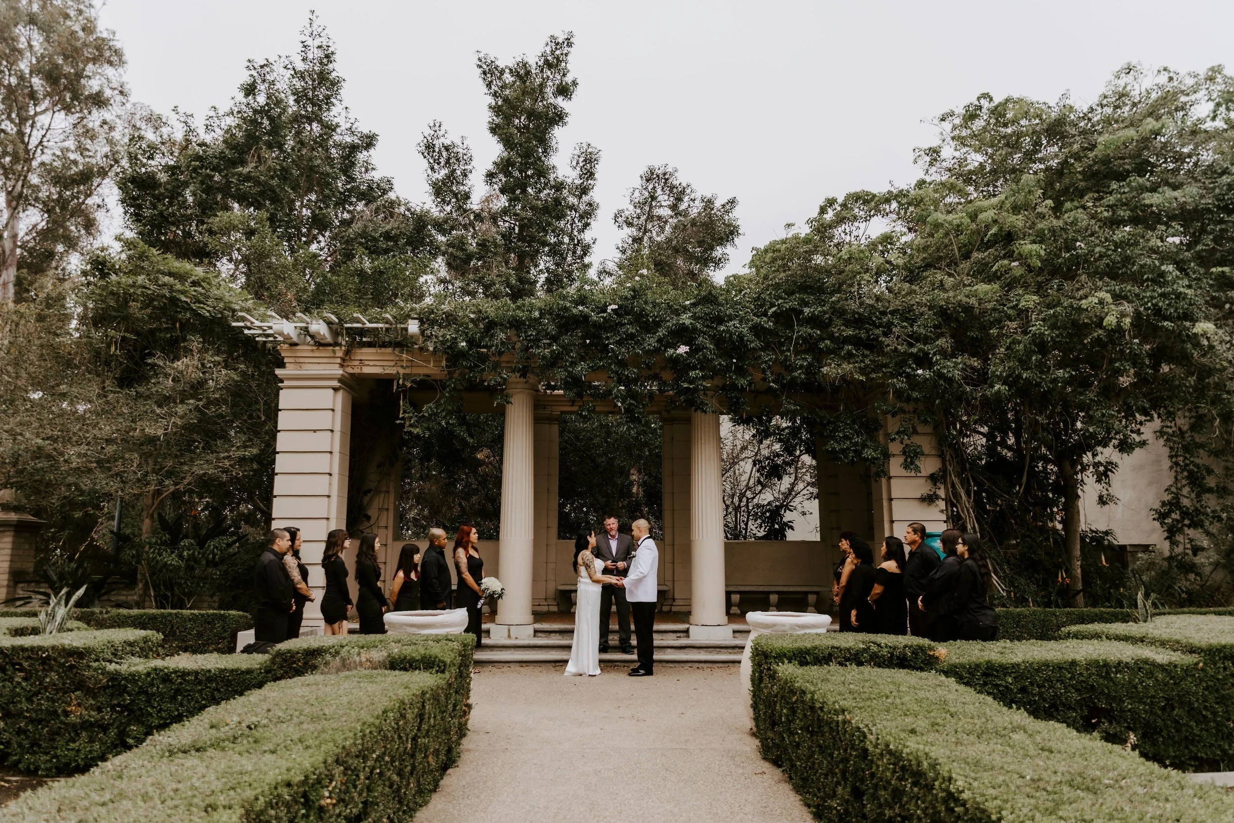 A wedding ceremony taking place outdoors under a classical gazebo with columns, surrounded by trees and manicured bushes. The bride and groom stand facing each other in the center, with an officiant, and guests are standing around watching.