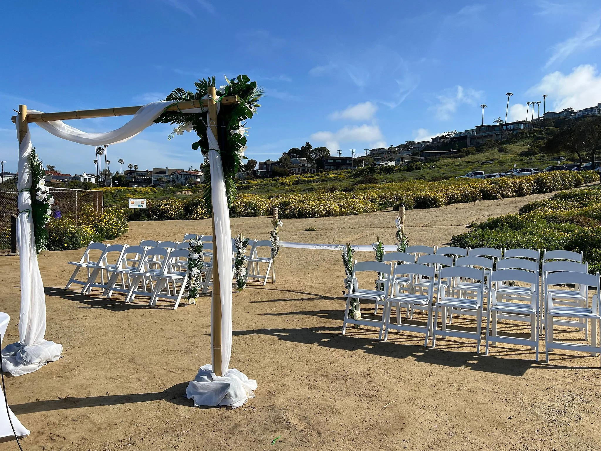 Wedding arch decorated with green leaves and white fabric, set on sandy ground with white chairs arranged for a ceremony, on a sunny day with houses and trees on a hillside in the background.