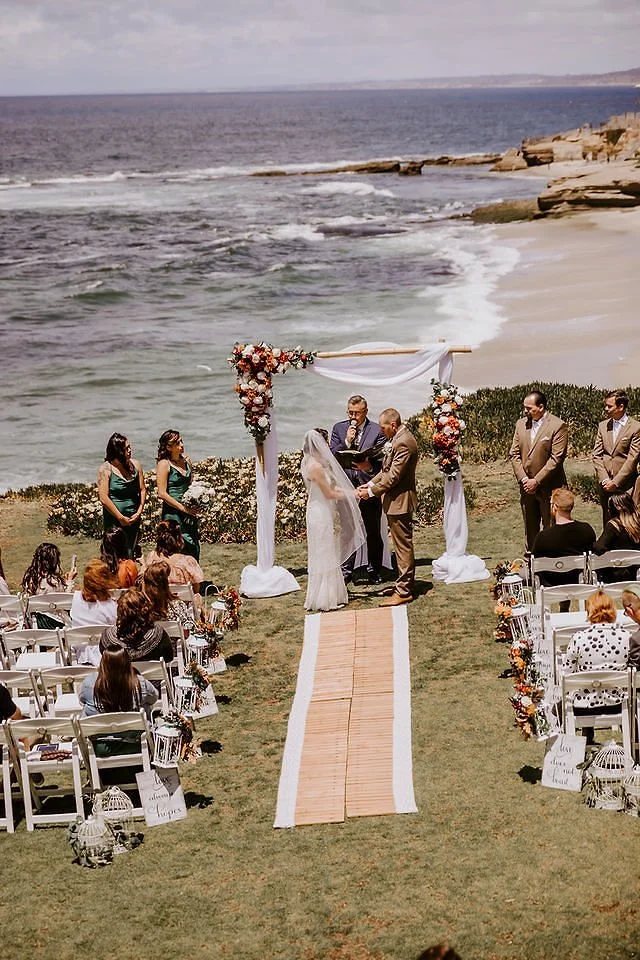 A wedding ceremony taking place on a grassy area near the beach with the ocean in the background. The bride and groom are holding hands under a decorated archway, with bridesmaids and groomsmen standing on either side. Guests are seated in white chai
