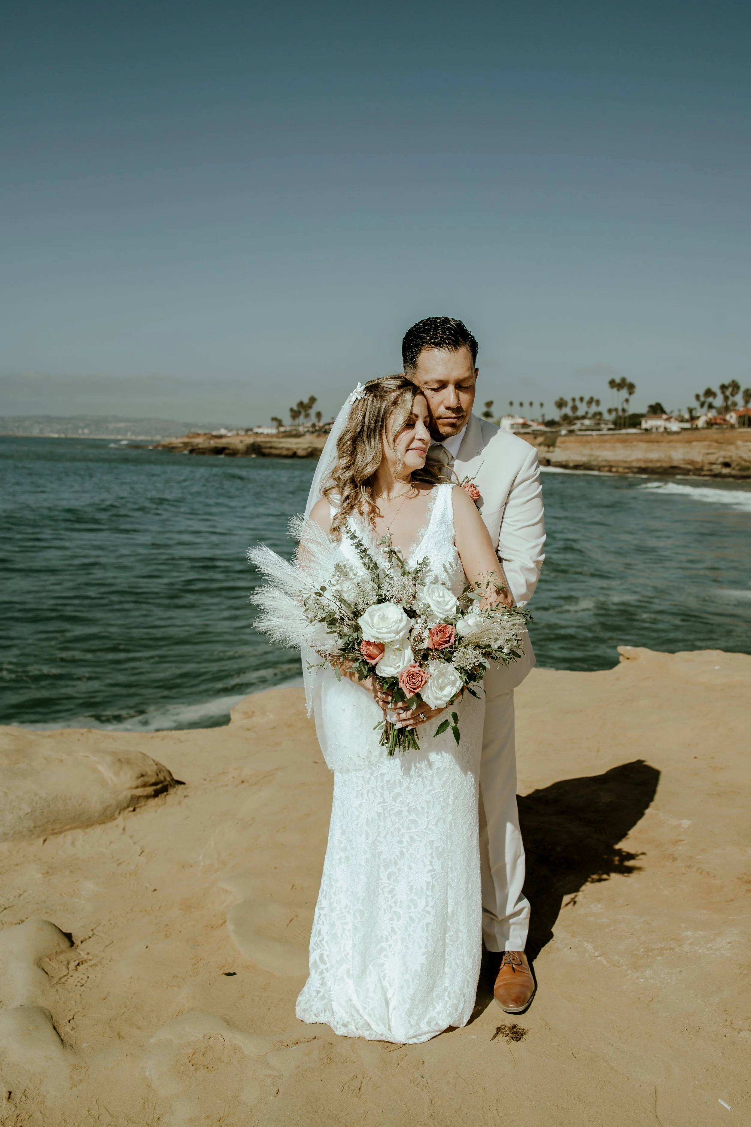 A bride and groom stand on a sandy beach, embracing each other. The bride holds a bouquet of white and pink roses with greenery, and the groom is dressed in a light-colored suit. The ocean and a rocky coastline with houses and palm trees are visible in the background, under a clear blue sky.
