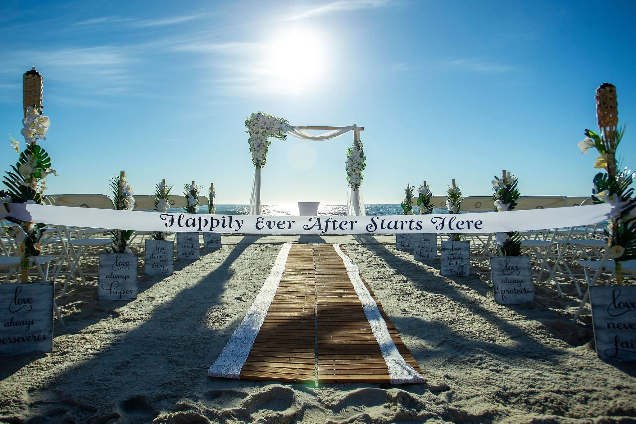 A wedding arch decorated with white flowers and white fabric on a platform near the ocean, with chairs and signs along a pathway, under a bright sun in a clear blue sky.