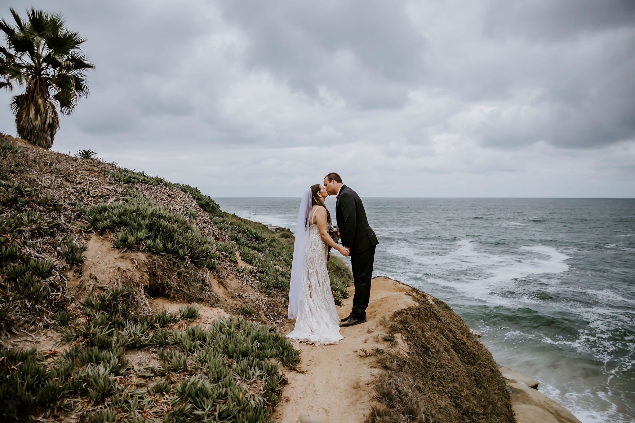 A bride and groom share a kiss on a rocky cliffside by the ocean under a cloudy sky, with lush green plants and a palm tree nearby.