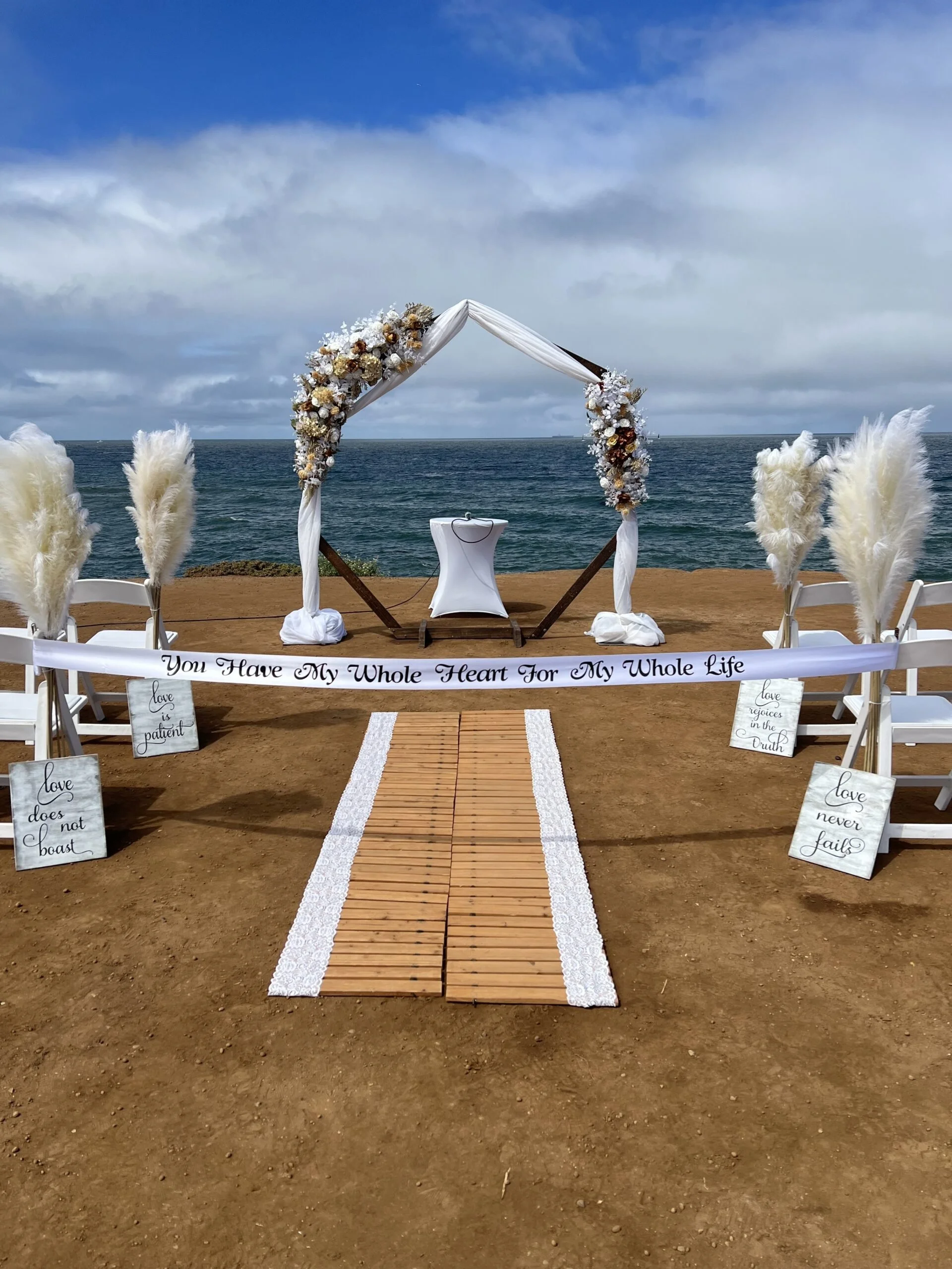 A wedding ceremony setup on a beach with a wooden aisle, white chairs, a floral arch, and ocean in the background. The arch is decorated with white and brown flowers and draped fabric. A small table is beneath the arch. There are signs with romantic quotes on the ground and a white banner with the text 'You Have My Whole Heart For My Whole Life'.