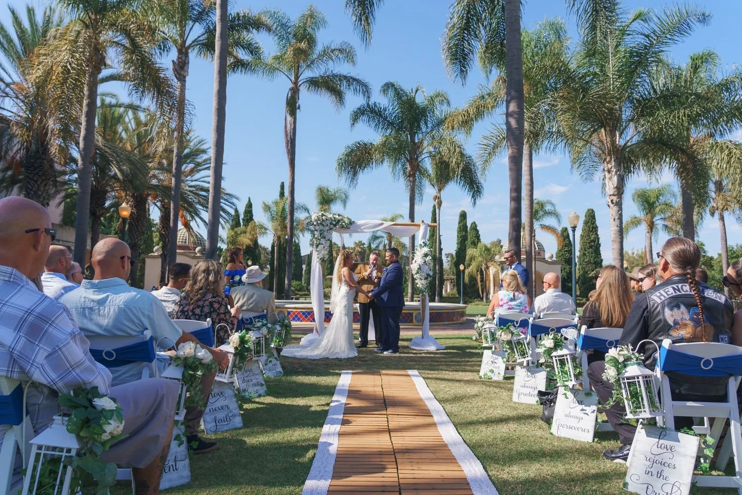 Outdoor wedding ceremony under tall palm trees with guests seated on either side and a couple exchanging vows at an arch decorated with white flowers.