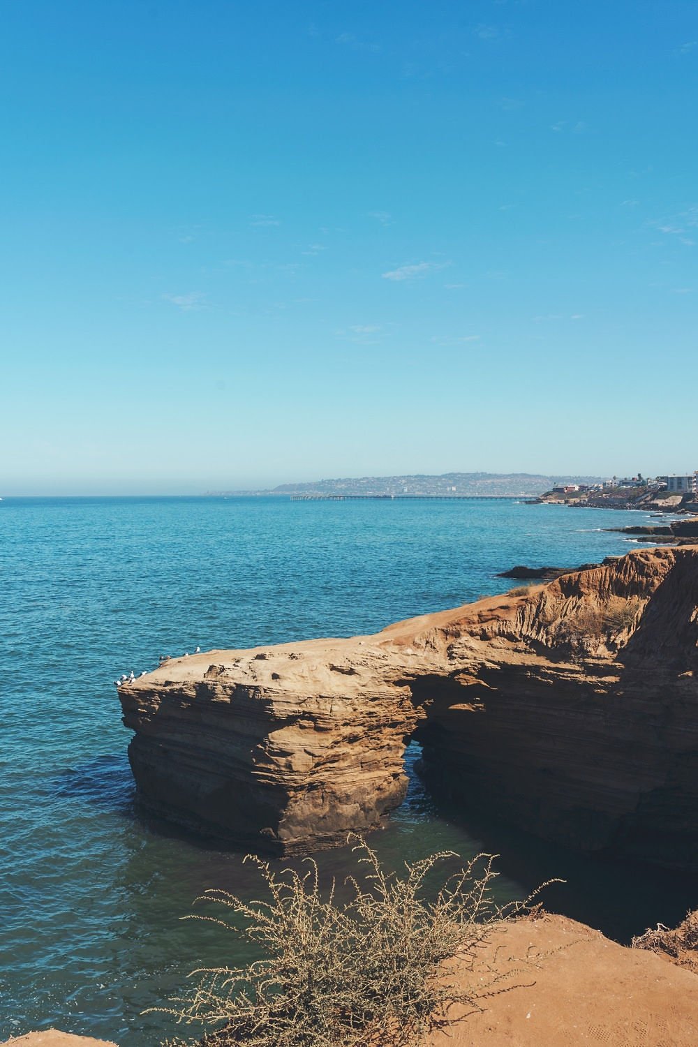 Coastal landscape with sandy cliffs and a natural rock arch over the ocean, under a clear blue sky.