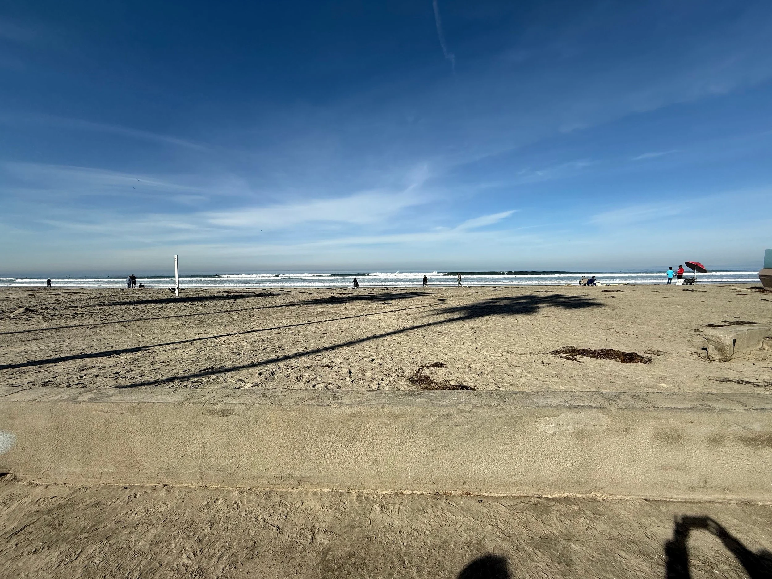 A sandy beach with footprints, a few people walking and sitting, a lifeguard stand, and a red umbrella with two people under it, with the ocean waves and a partly cloudy sky in the background.