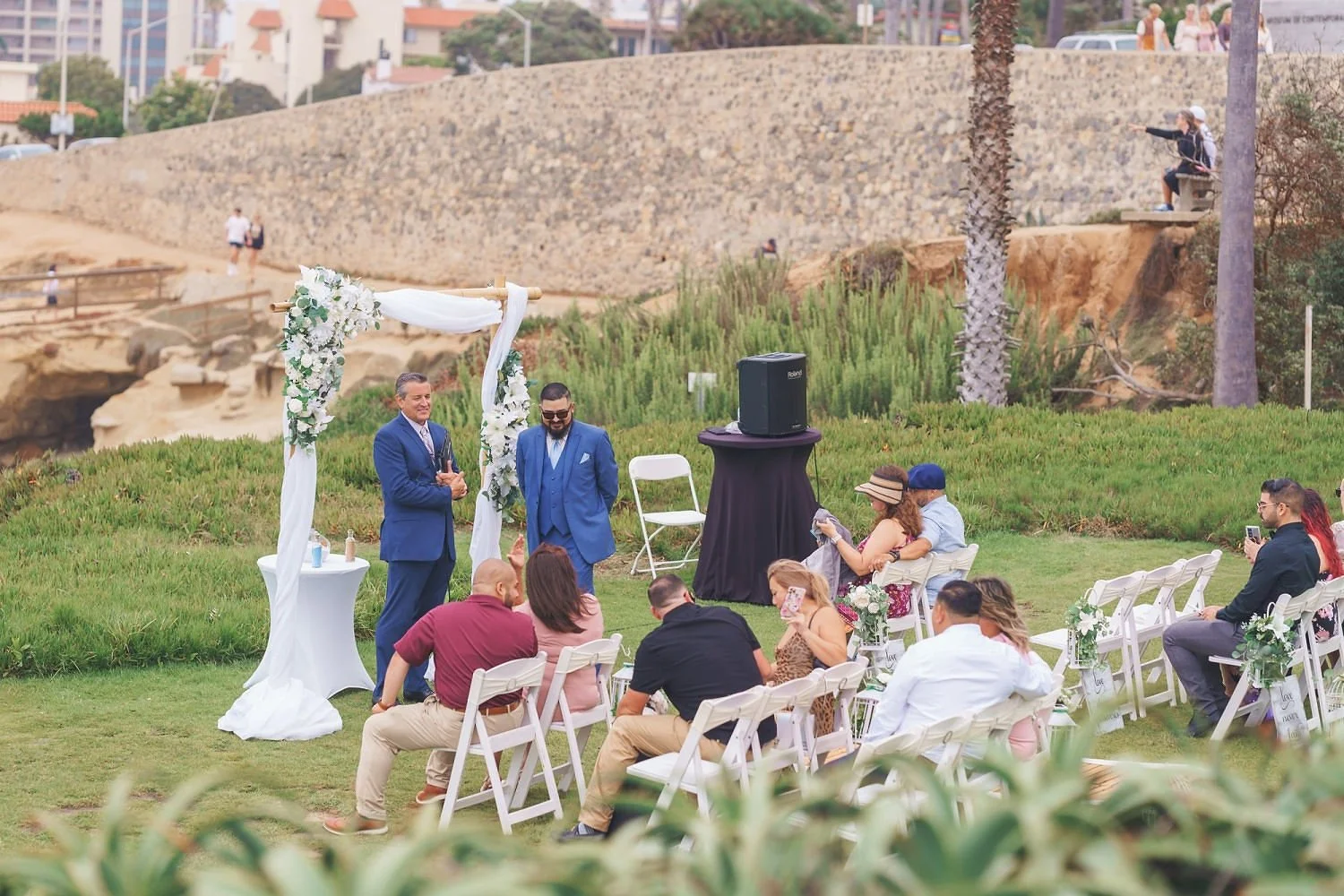 Outdoor wedding ceremony with a couple standing under a floral arch, guests seated on white chairs, some taking photos, in a grassy area with trees and a stone wall in the background.