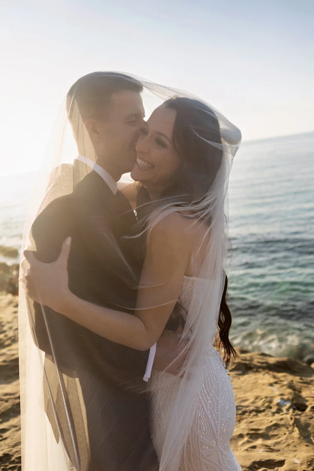 A bride and groom hugging on a beach, with the ocean and sky in the background, during sunset.