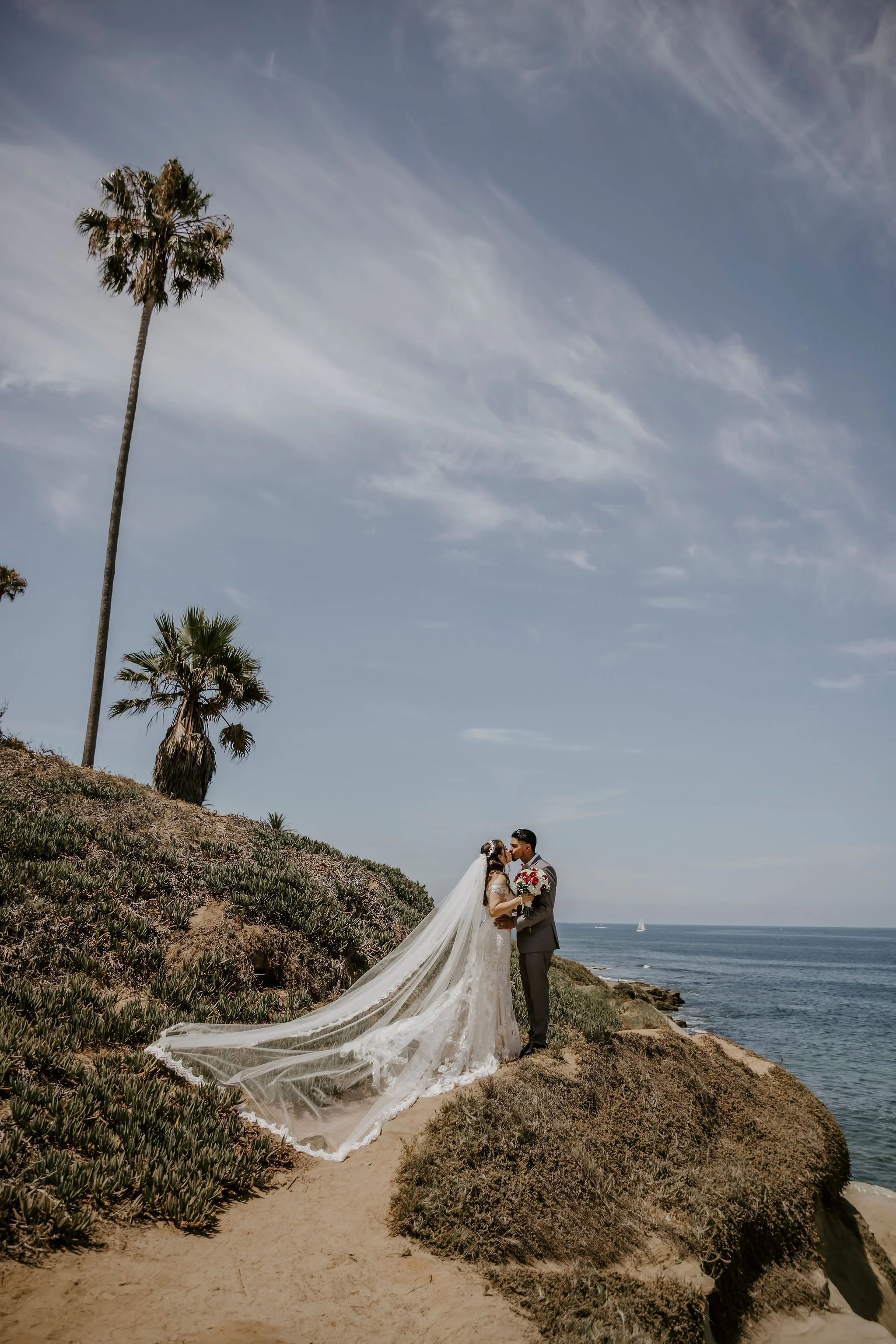 A bride and groom standing on a rocky, grassy hillside by the coast, with palm trees and the ocean in the background. The bride wears a long white wedding gown with a veil, and the groom wears a gray suit. They are holding each other and sharing a mo