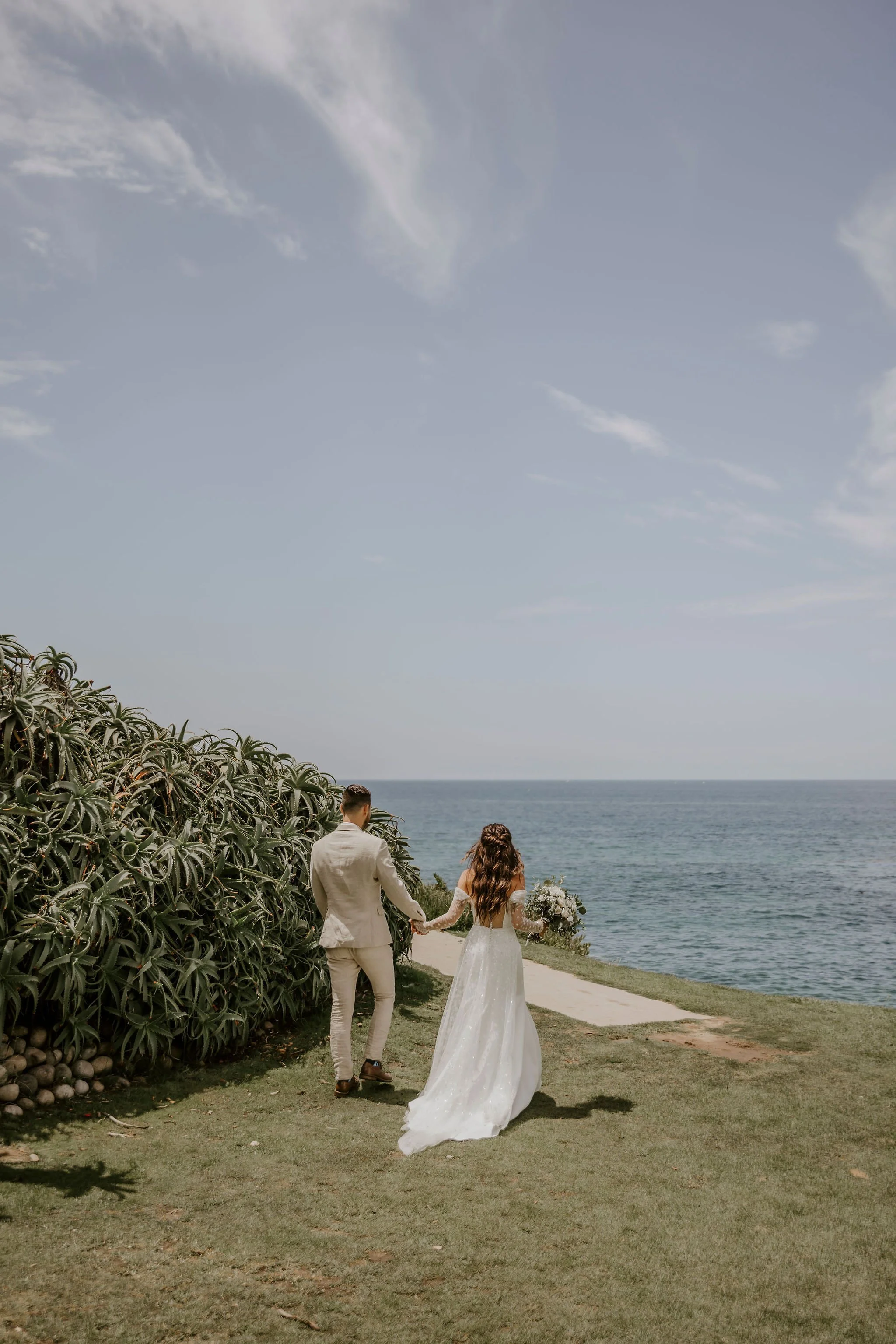 A wedding couple walking hand in hand along a path near the ocean. The woman is in a white wedding dress holding a bouquet, and the man is in a light-colored suit. They are surrounded by greenery, with the ocean and sky in the background.
