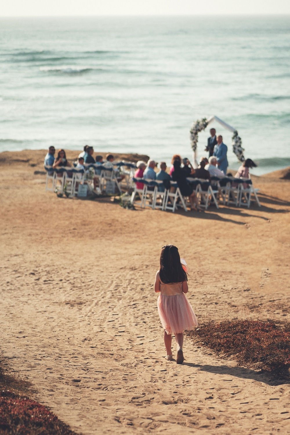 A young girl walking on a sandy path towards a wedding ceremony set up on a beach with the ocean in the background.