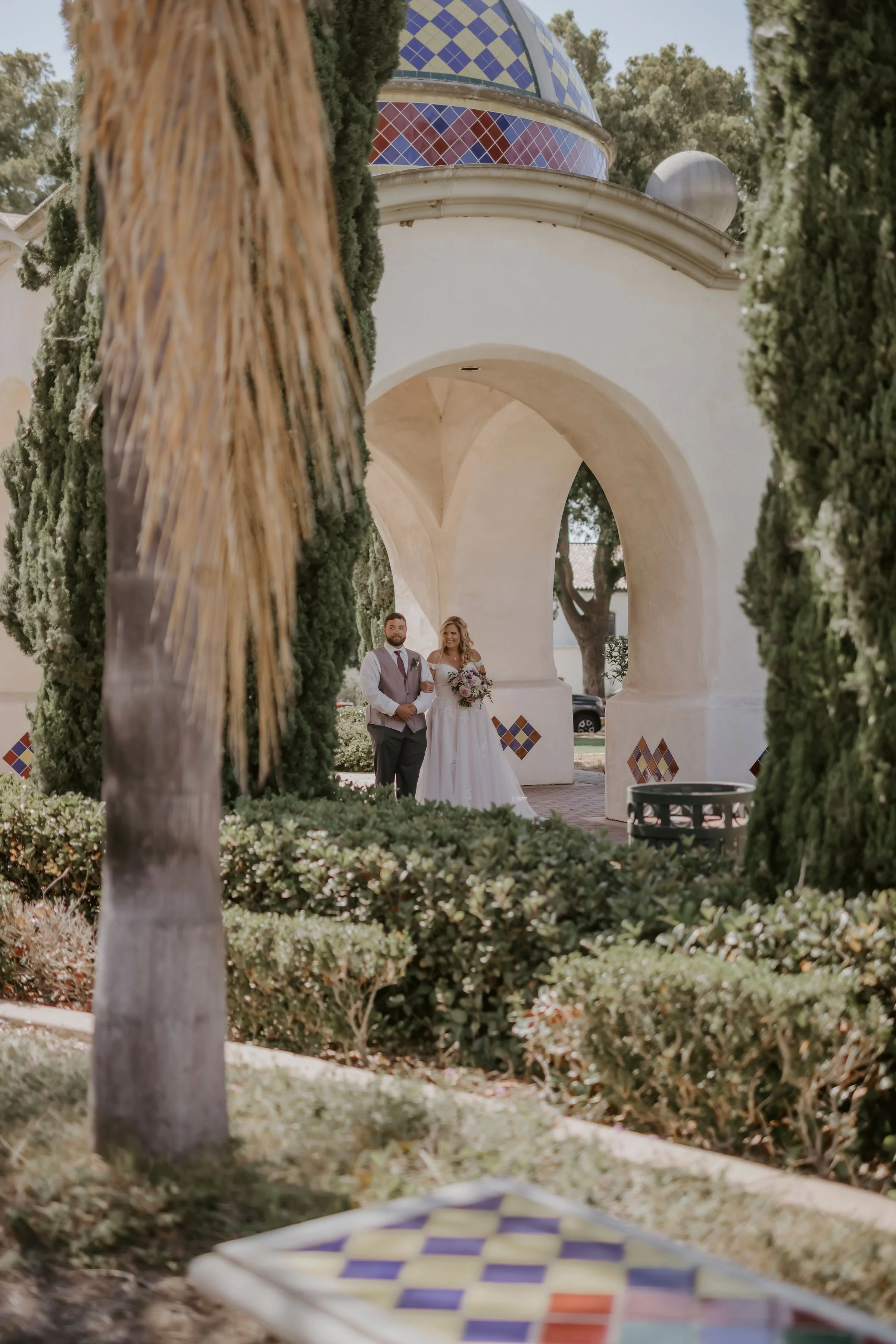 A bride and groom standing together under an archway, surrounded by trees and bushes in an outdoor setting. The bride is holding a bouquet, and both are dressed in formal wedding attire.