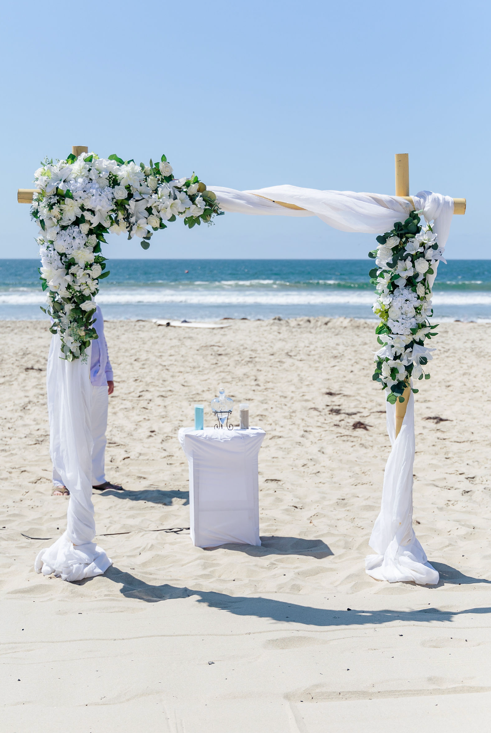 A wedding altar decorated with white flowers and fabric on a sandy beach with the ocean in the background.