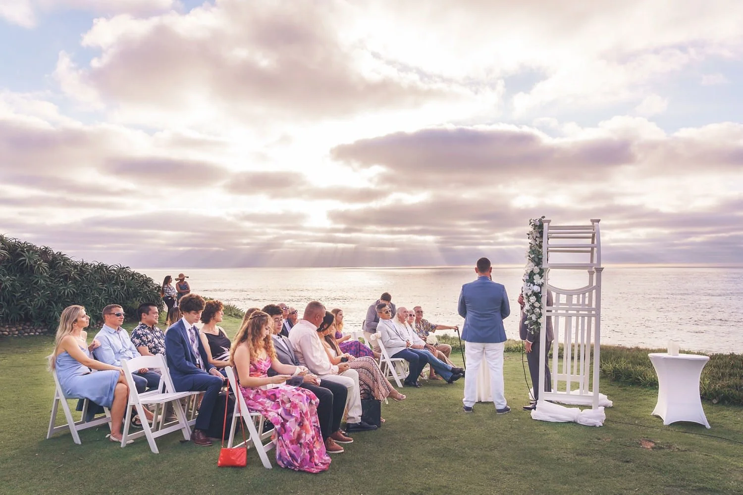 A wedding ceremony taking place outdoors near the ocean during sunset, with guests seated on white chairs facing an officiant and groom.