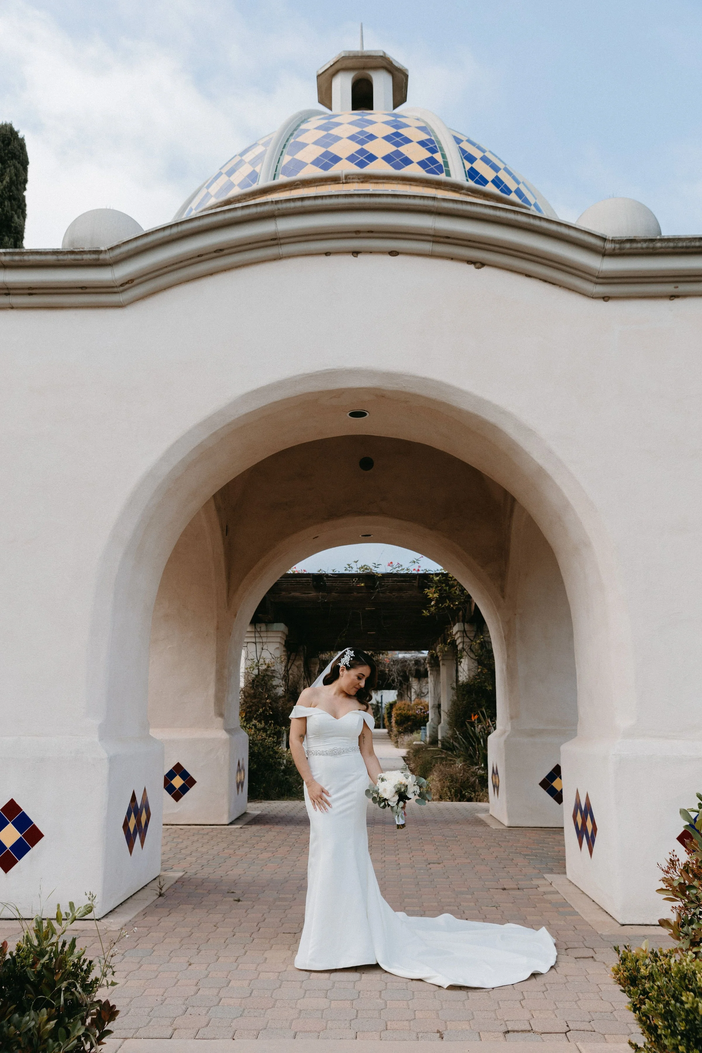 A bride in a white wedding dress holding a bouquet, standing under a white archway with a blue-tiled dome in the background, during daytime.