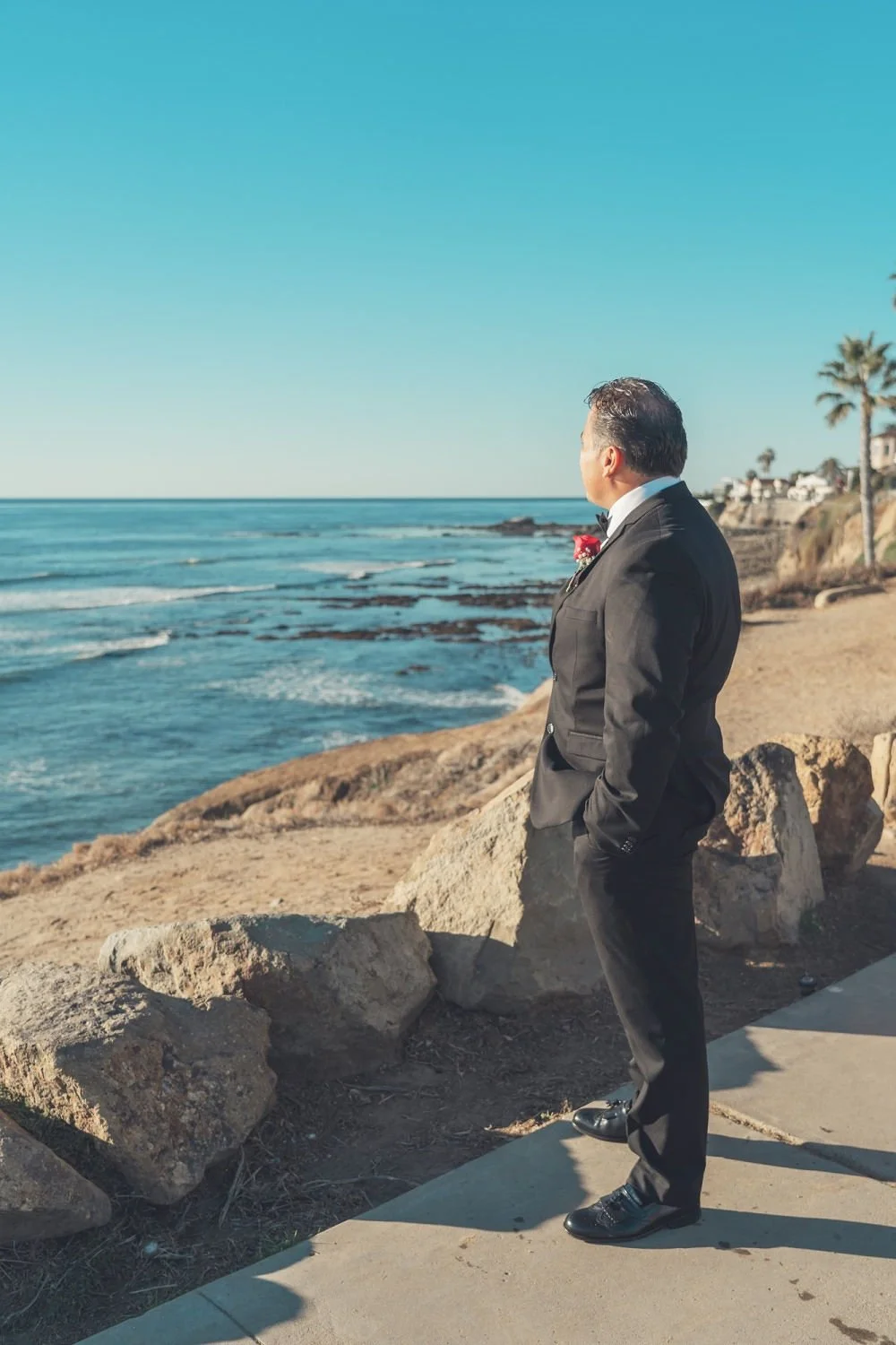 A man in a tuxedo stands on a sidewalk near the beach, looking out over the ocean with palm trees and coastal houses in the background.