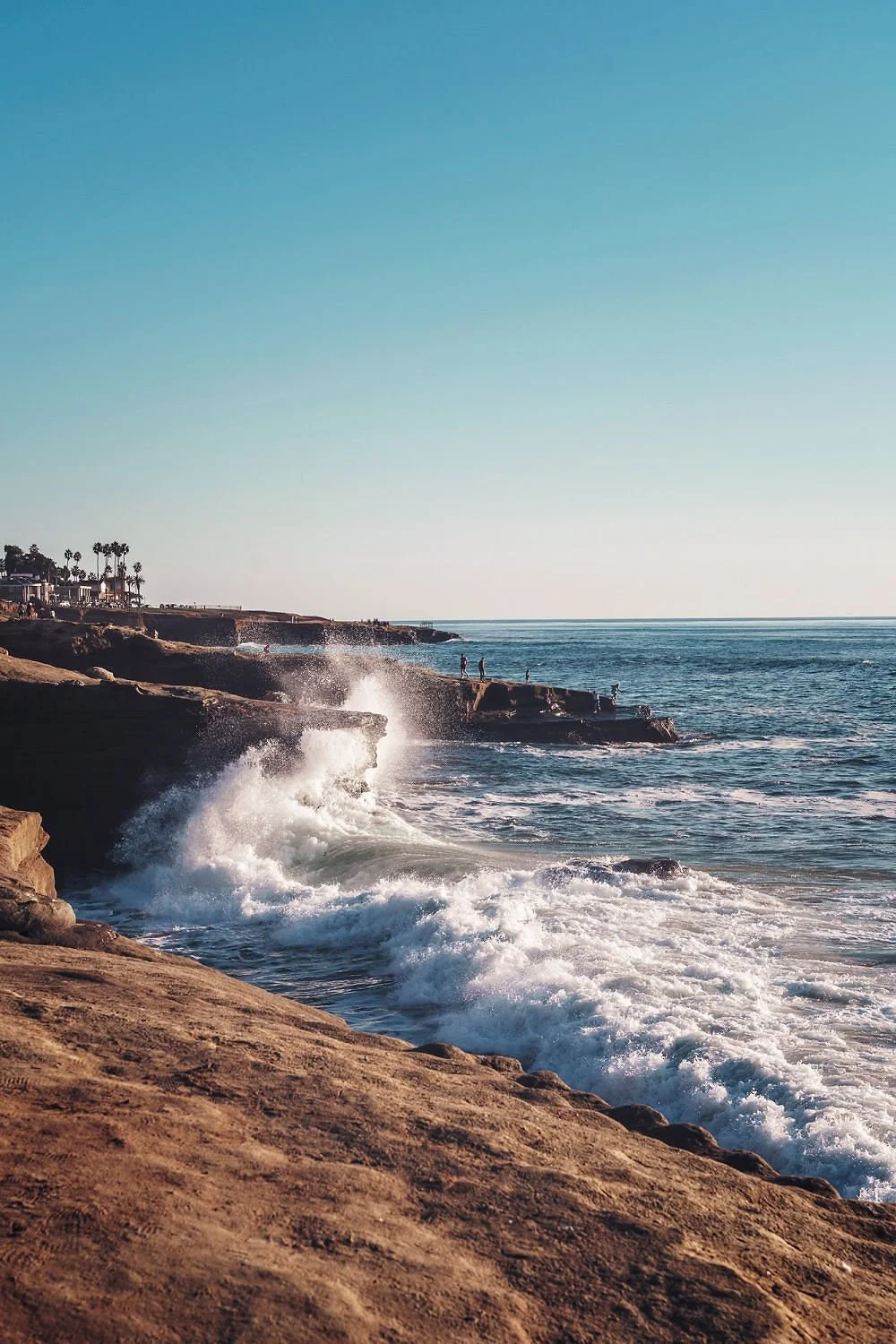 Coastal scene with rocky cliffs, ocean waves crashing against rocks, a few people walking on the cliffs, and palm trees in the background under a clear blue sky.