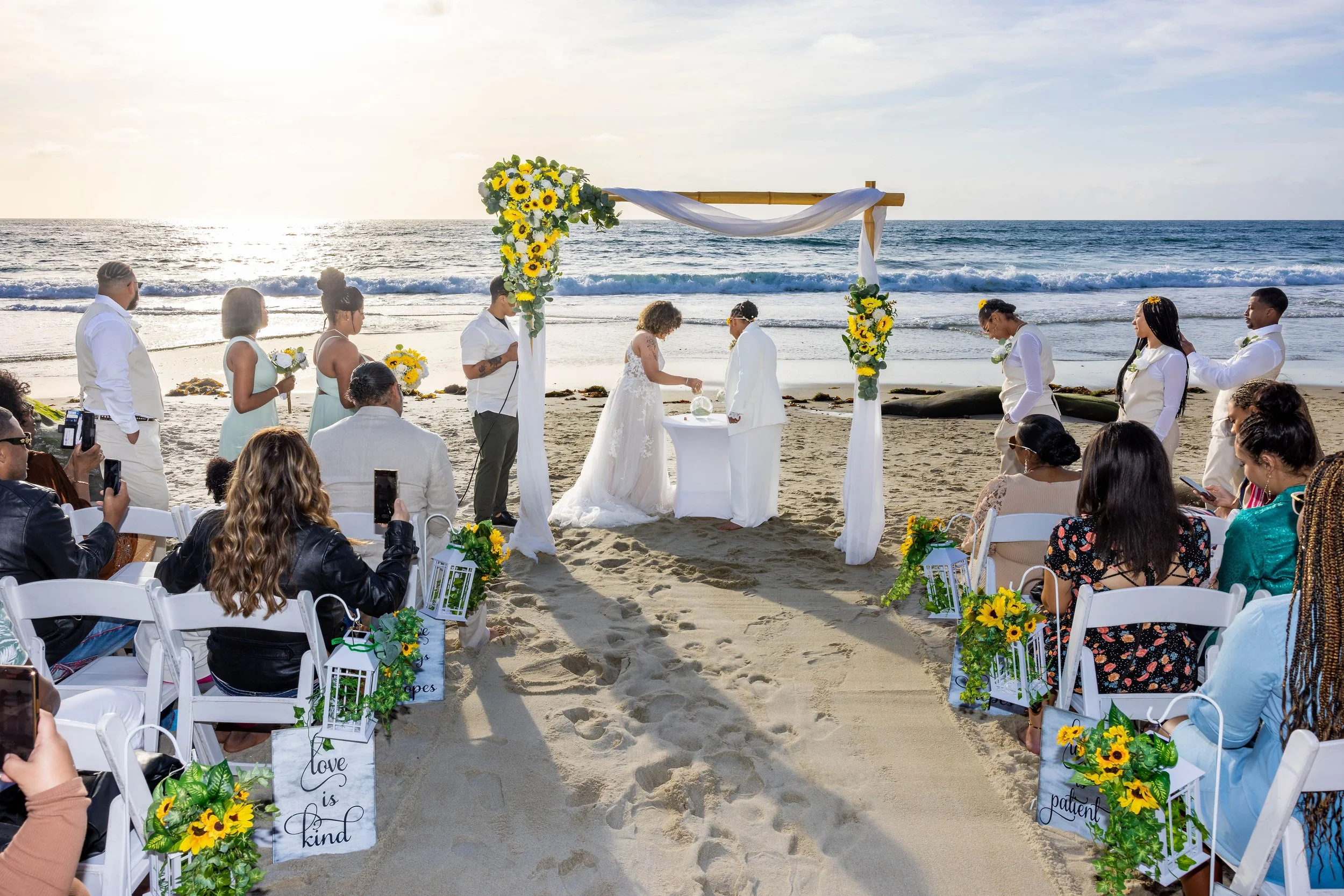 A beach wedding ceremony with a floral arch, seated guests, and a couple exchanging vows near the ocean at sunset.