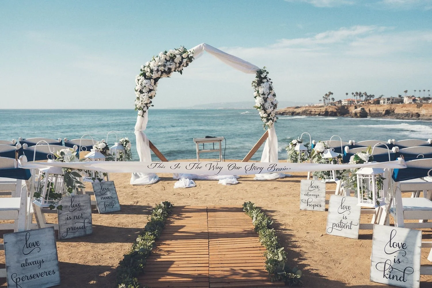 Beach wedding setup with a floral arch, wooden aisle, and chairs with decorative signs, ocean and coastline in the background.