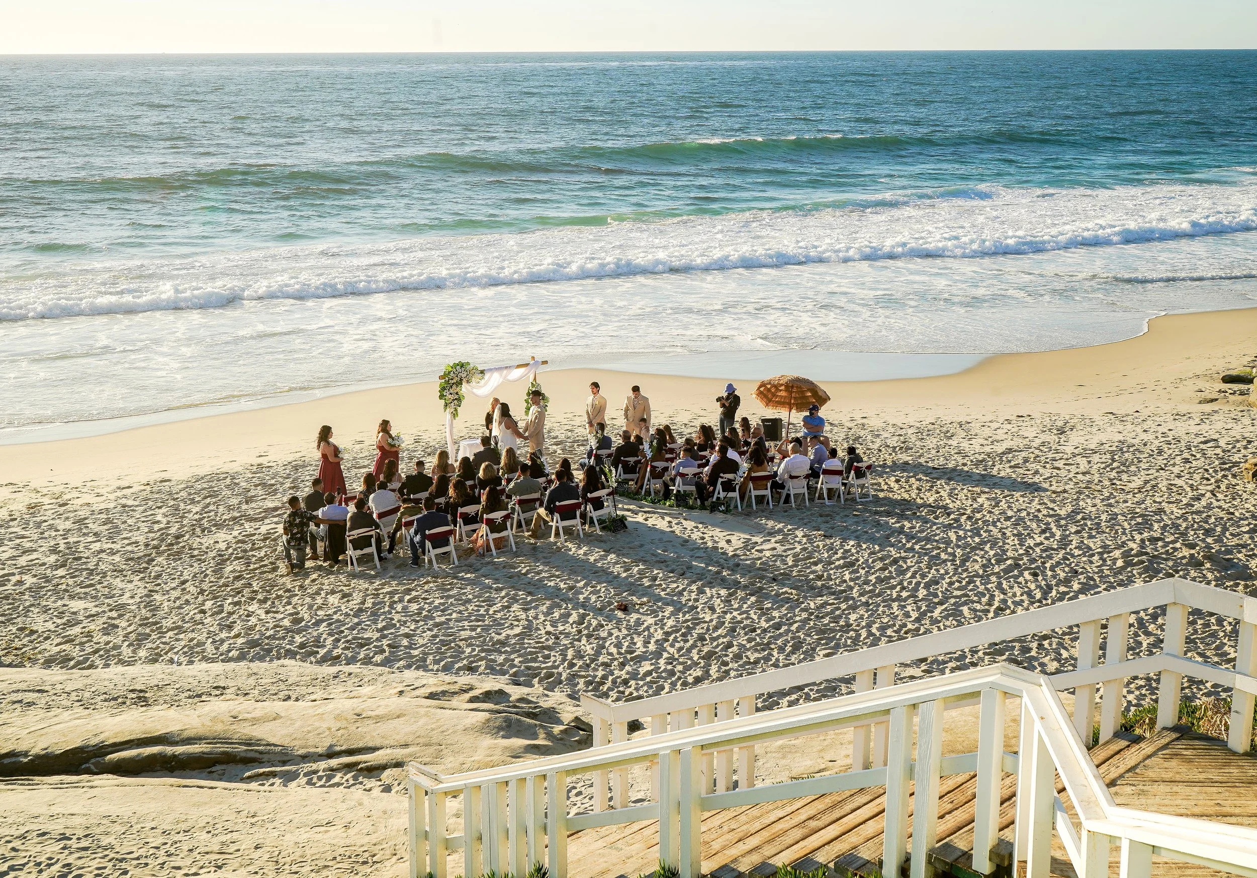 A beach wedding ceremony taking place on sandy shore, with guests seated in rows and a couple exchanging vows under a decorated arch, ocean waves in the background.