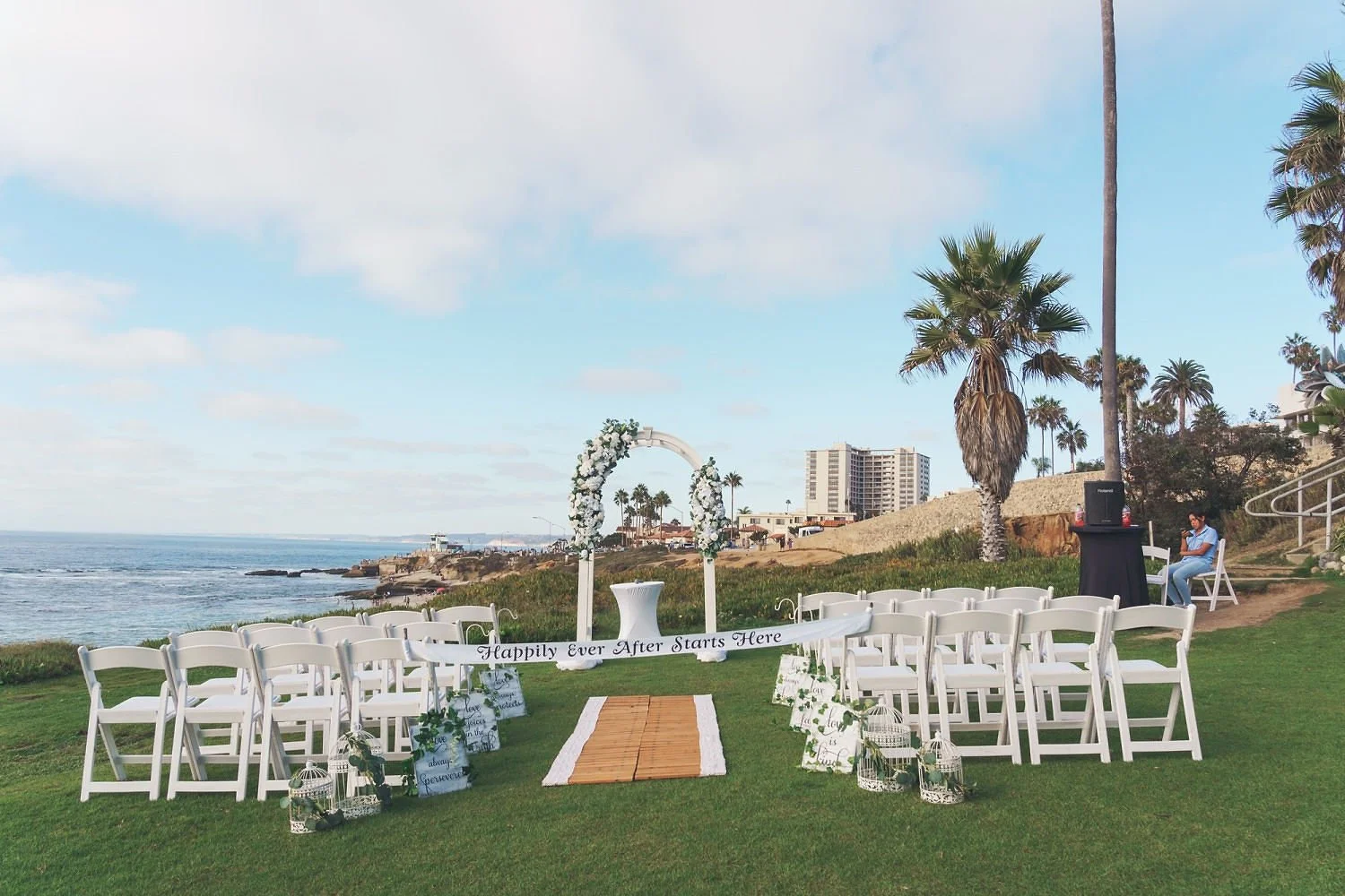 Beachside wedding setup with white chairs, an arch decorated with white flowers, a banner with the saying "Happily Ever After Starts Here," and a wooden aisle on green grass next to the ocean, with palm trees and buildings in the background.