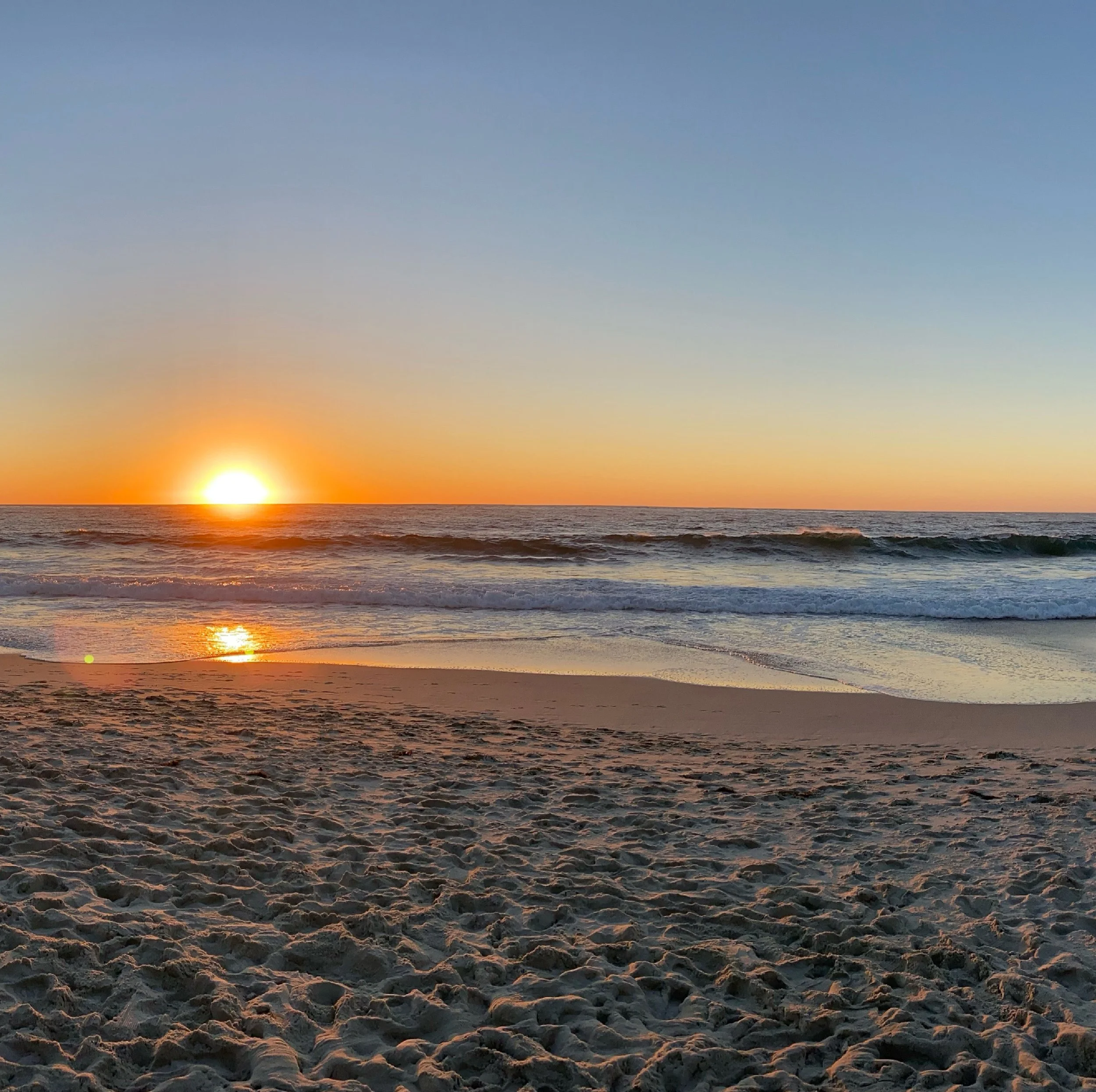 Sunset over the ocean with sandy beach foreground, gentle waves, and colorful sky.