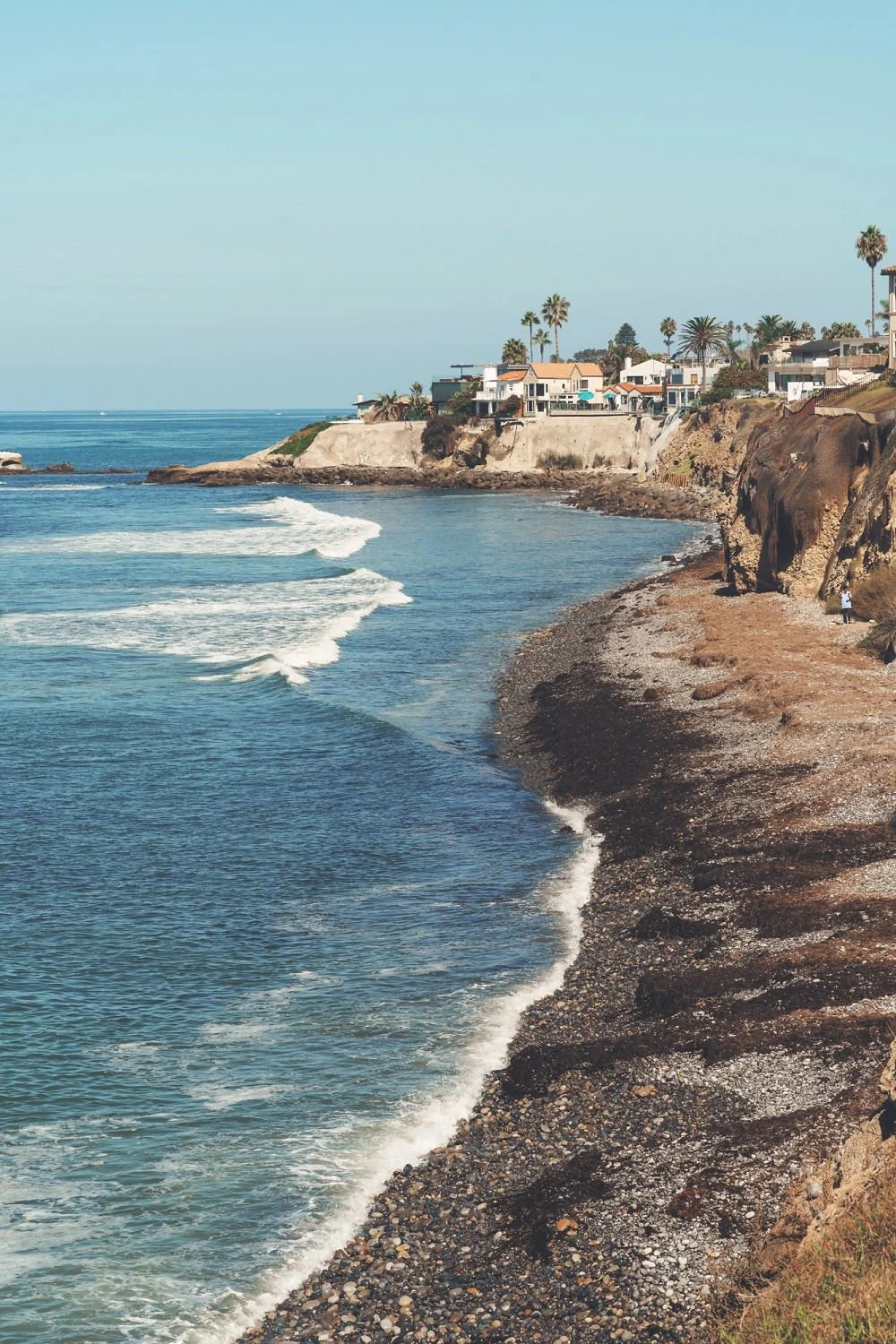 A coastal scene featuring a rocky shoreline with waves crashing, cliffside houses, and palm trees against a clear blue sky.