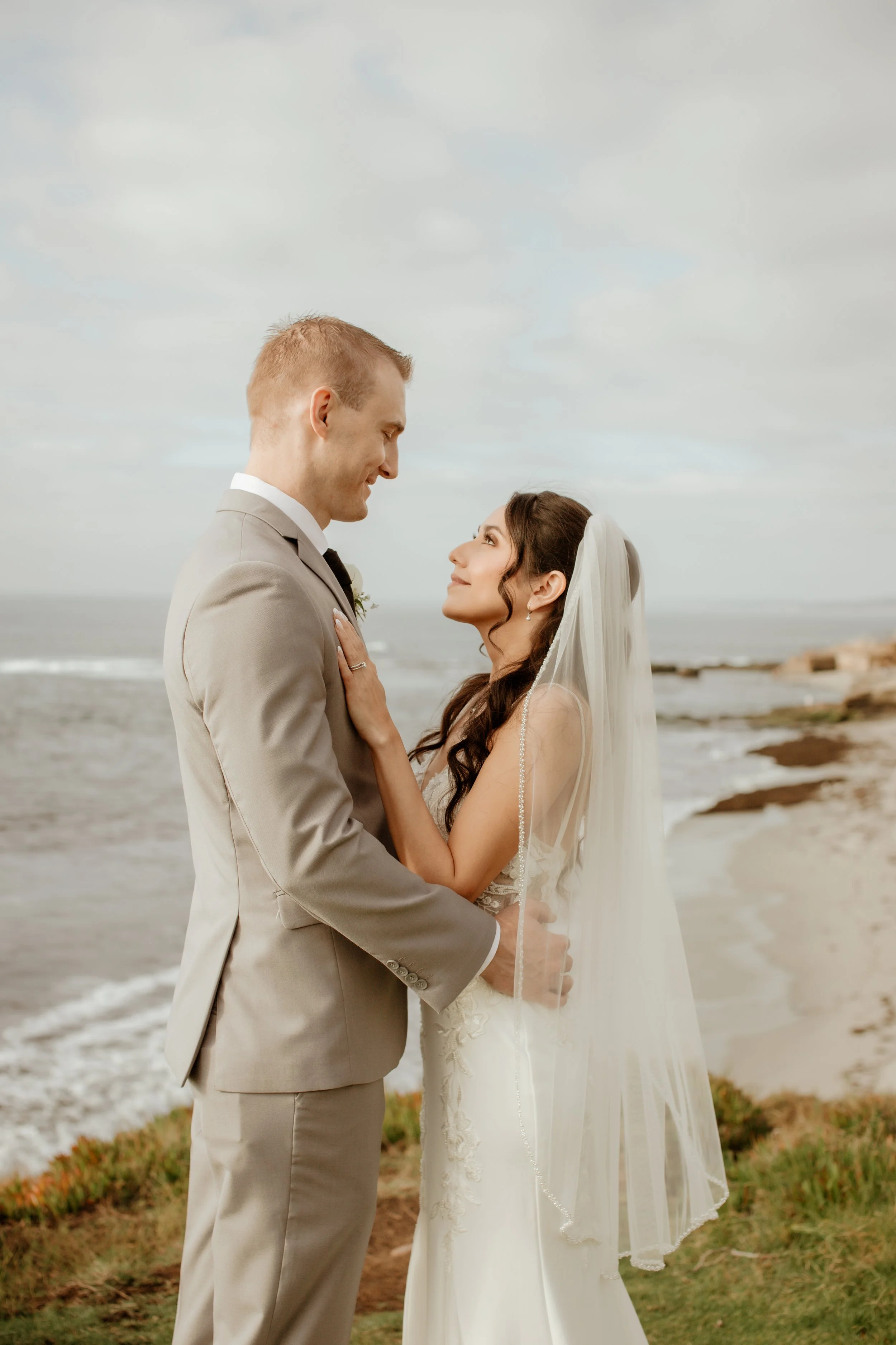 A bride and groom in wedding attire standing on a beach, gazing into each other's eyes, with the ocean and cloudy sky in the background.