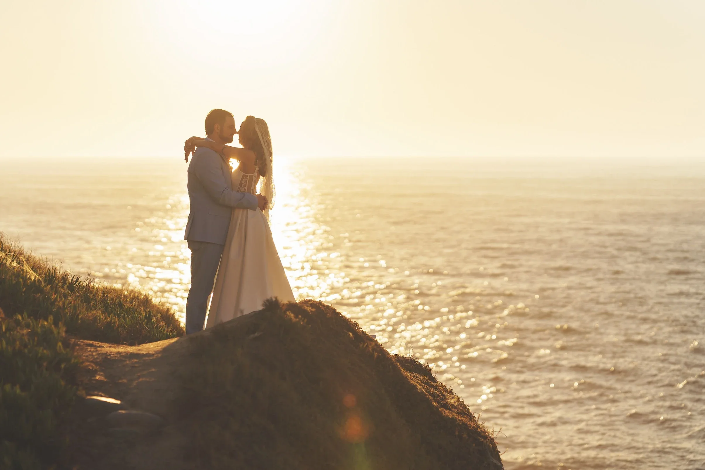 A couple dressed in wedding attire sharing a kiss at sunset on a seaside cliff, with the ocean in the background.