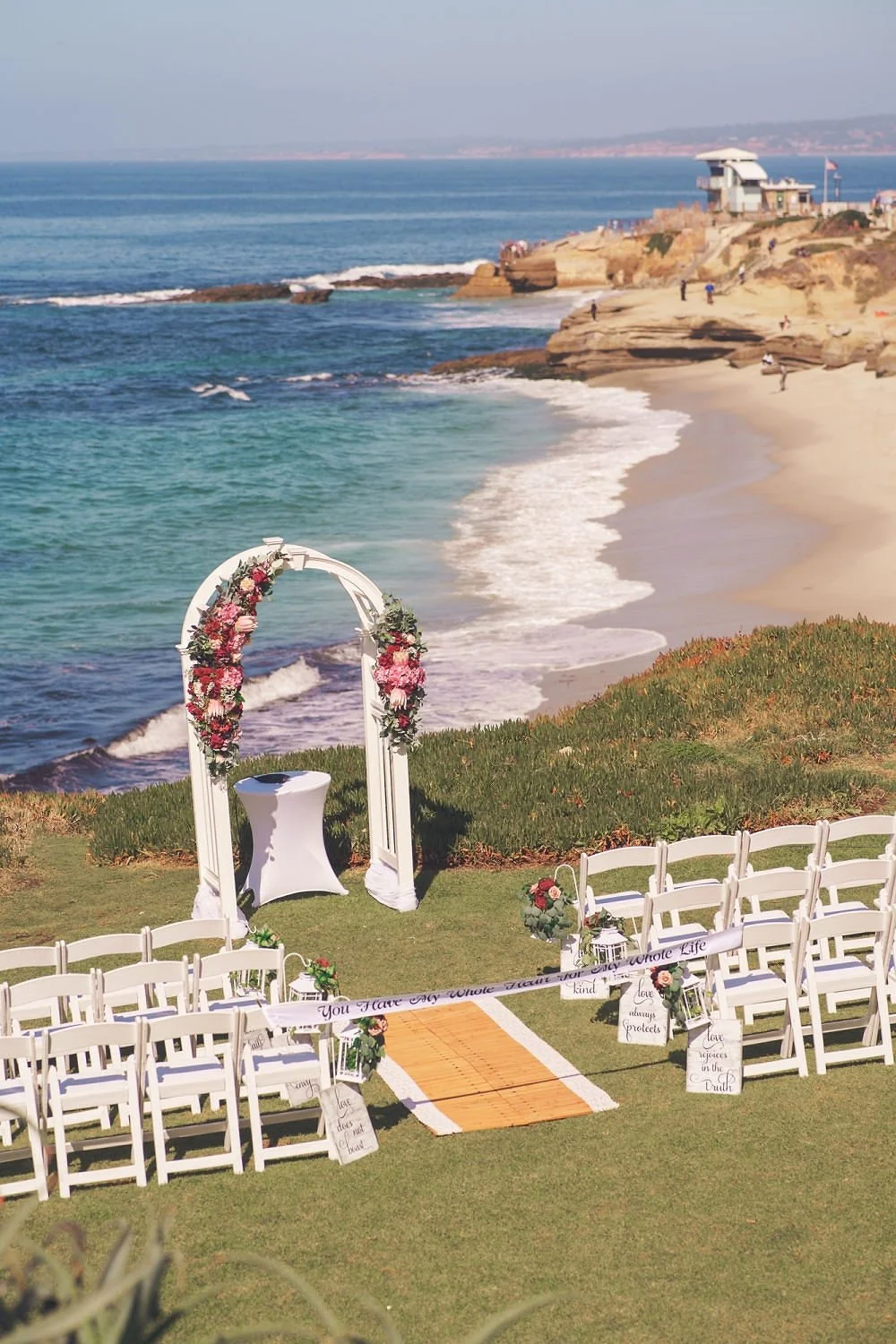 Outdoor wedding setup on a grassy area overlooking the ocean with white chairs, floral decorations, and an archway with flowers.