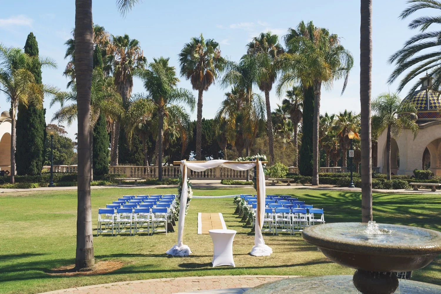 Outdoor wedding setup in a lush garden with palm trees, white chairs, an arch with white fabric, and a pathway, under a clear blue sky.
