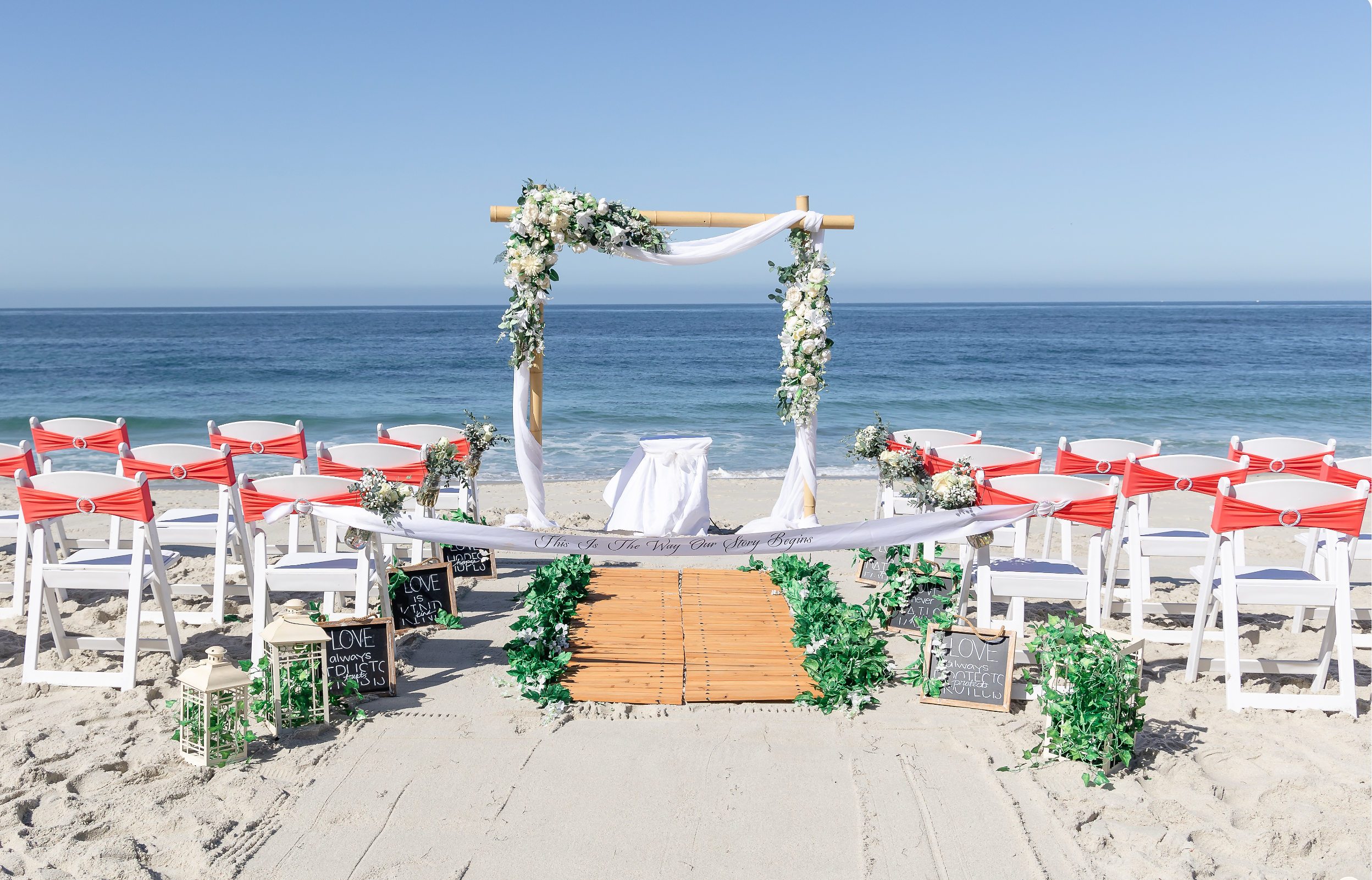 Beach wedding setup with white chairs adorned with red sashes, a wooden aisle with greenery, and a floral arch on the sand overlooking the ocean.