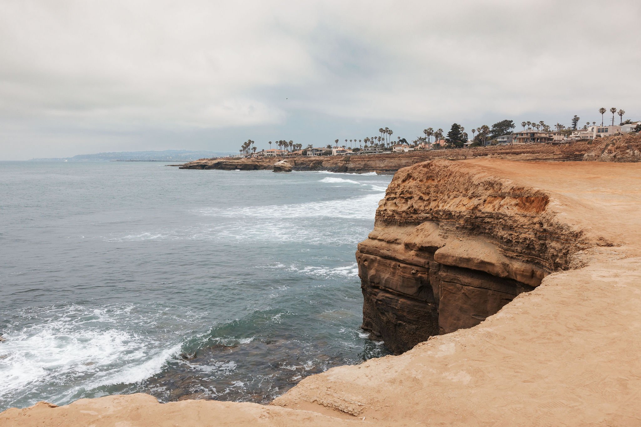 Cliffs along a cloudy coastline with houses and palm trees on top, and ocean waves crashing below.