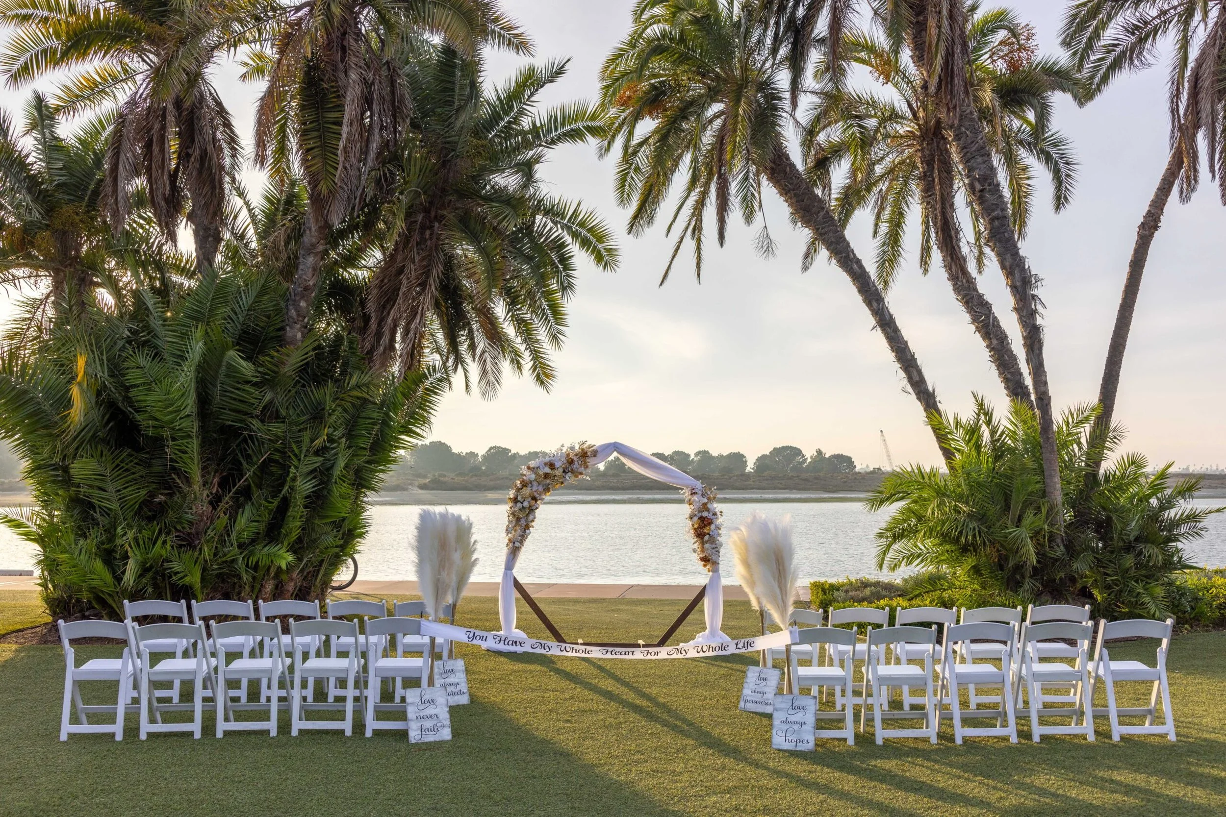 Outdoor wedding ceremony setup near a river with white chairs, an arch decorated with flowers, and tall palm trees in the background.