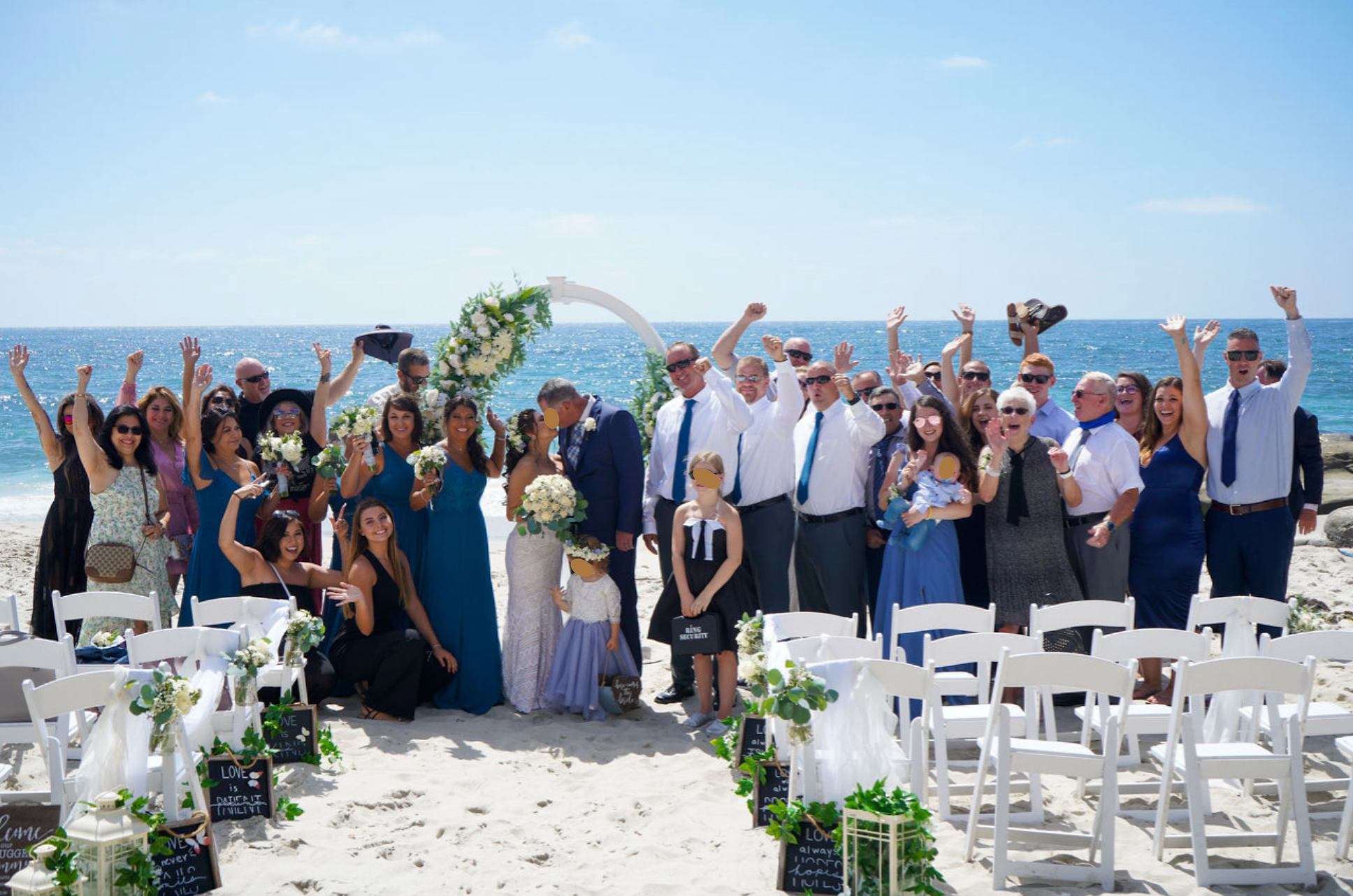 A large group of people gathered at the beach for a wedding, with the ocean and a blue sky in the background, and an arch decorated with flowers.