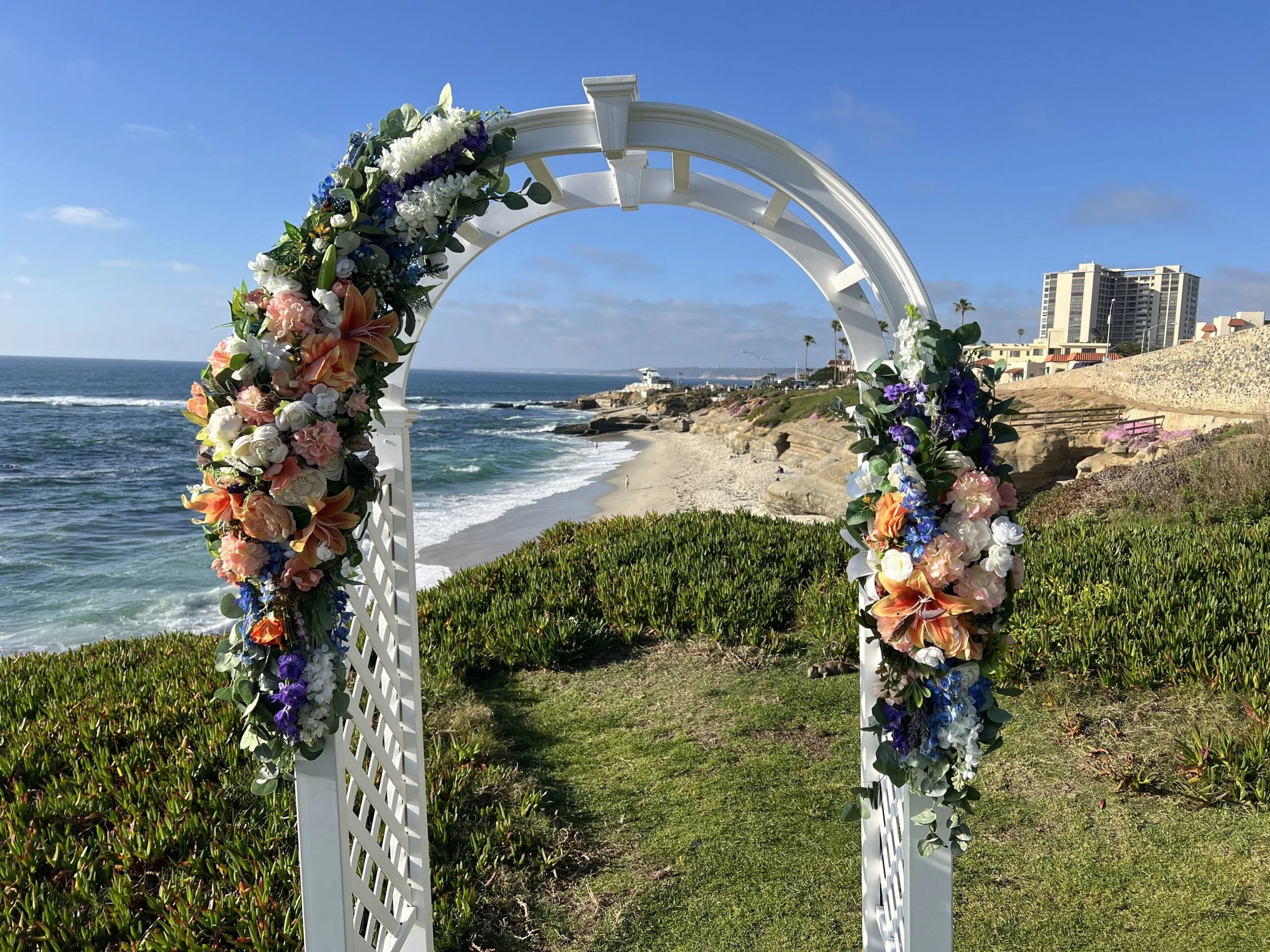 A white decorative arch decorated with colorful flowers, situated on a grassy area by the beach with the ocean and buildings in the background.
